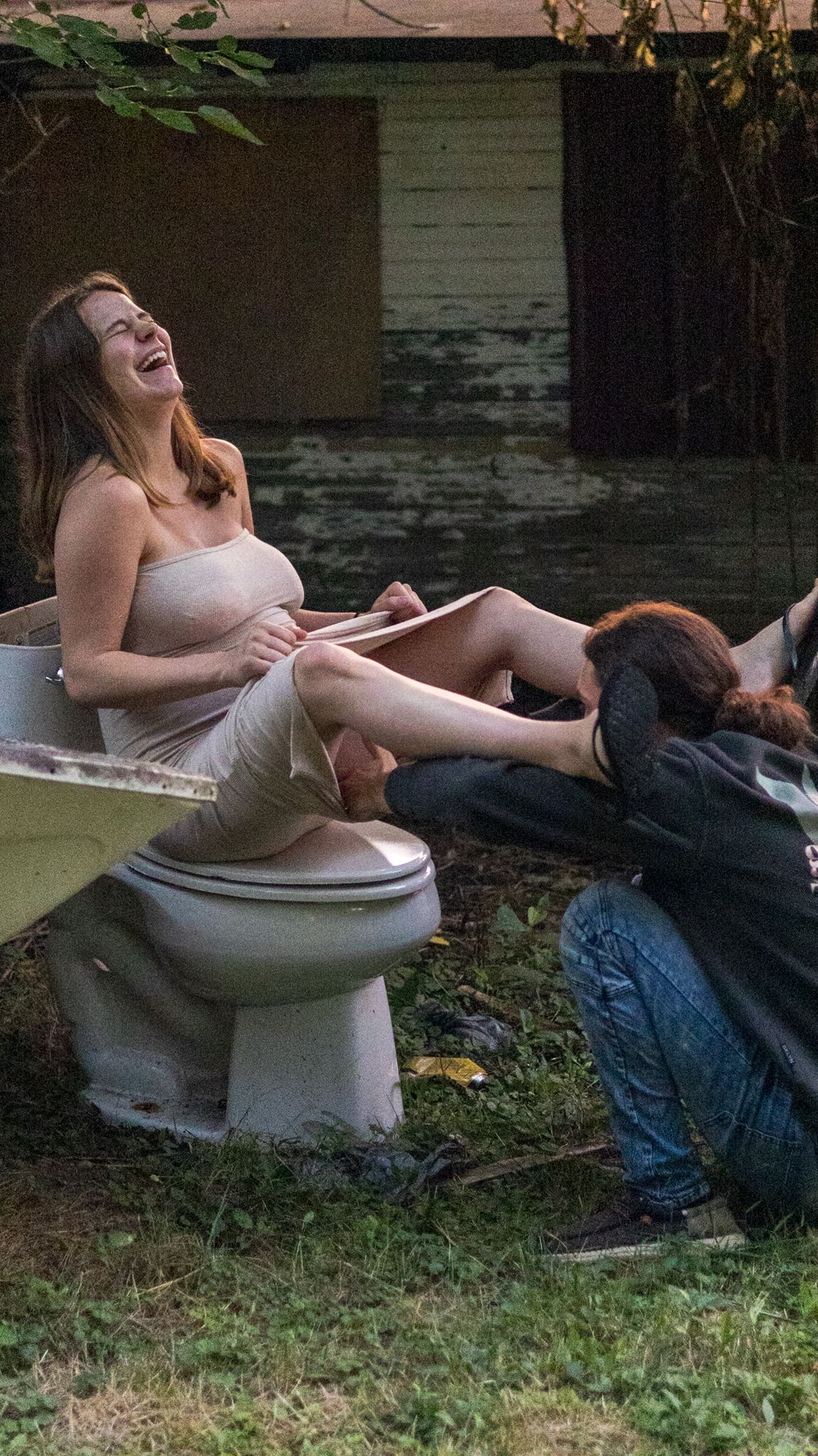 Two women laughing outdoors near an old bathtub and toilet in an overgrown backyard.