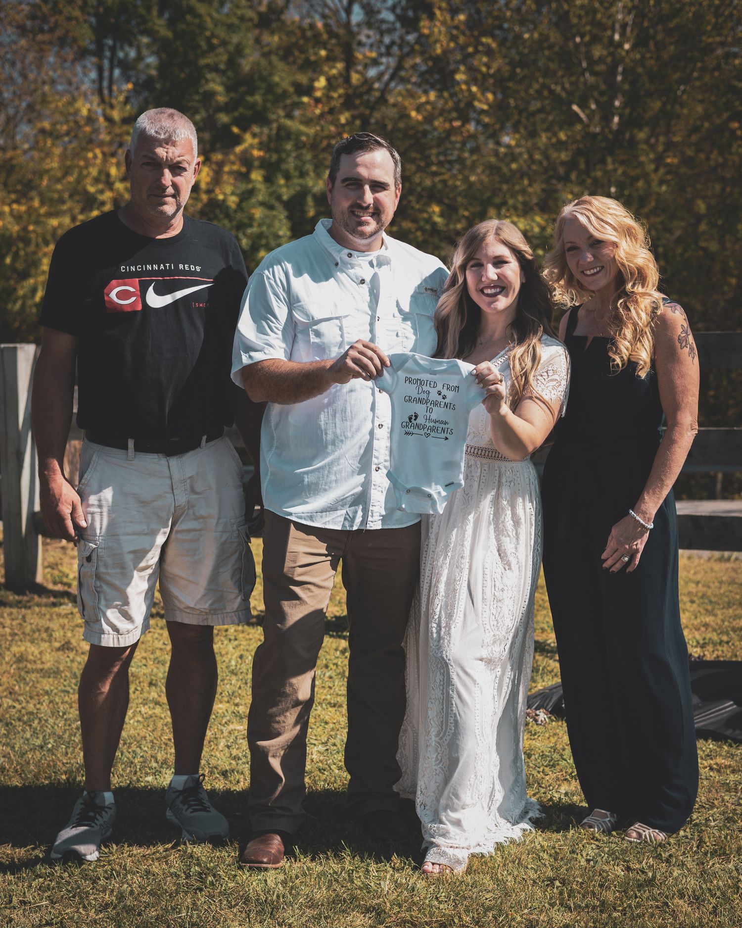 Family of four poses together holding a baby onesie at an outdoor gender reveal party on a sunny day.