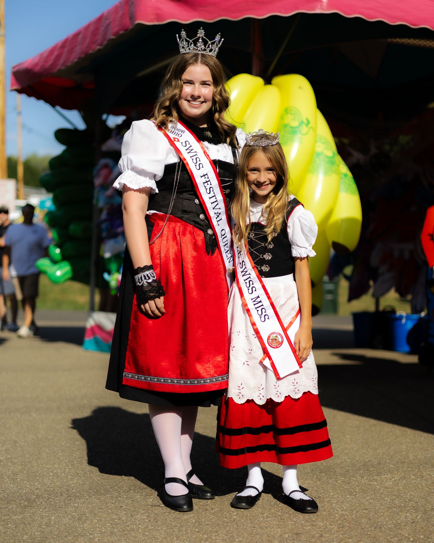 Two girls in traditional Swiss festival dresses and crowns posing at outdoor fair.