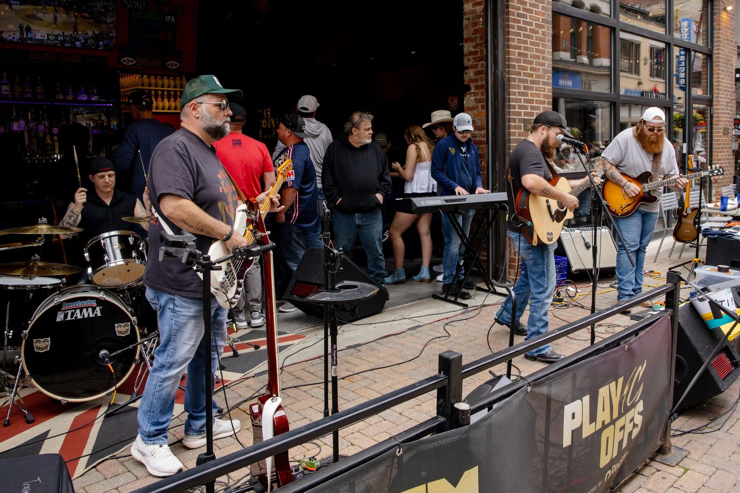 Live band performing outdoors on a sidewalk stage near a bar, with musicians playing guitars and drums for a crowd.