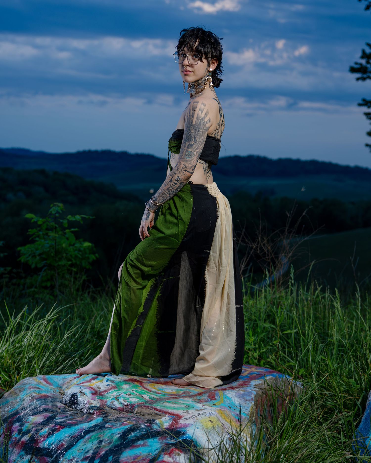 Tattooed woman in green and black patchwork skirt stands on painted rock at dusk with rolling hills behind.