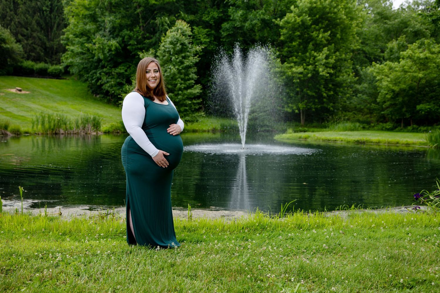 Smiling pregnant woman in green maxi dress posing by serene pond with water fountain backdrop.