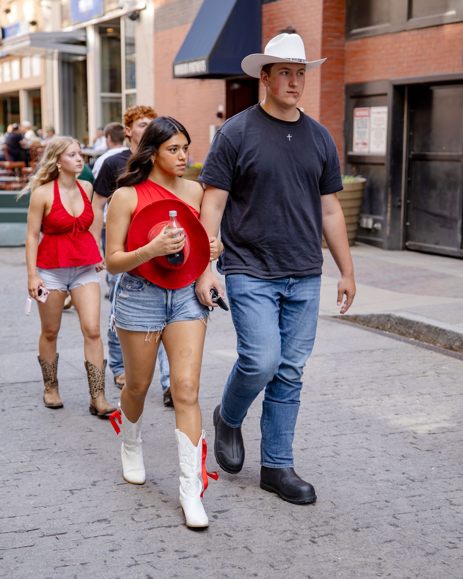 Two people in cowboy hats and boots walk a city street, woman in red top and denim shorts holds a red hat.