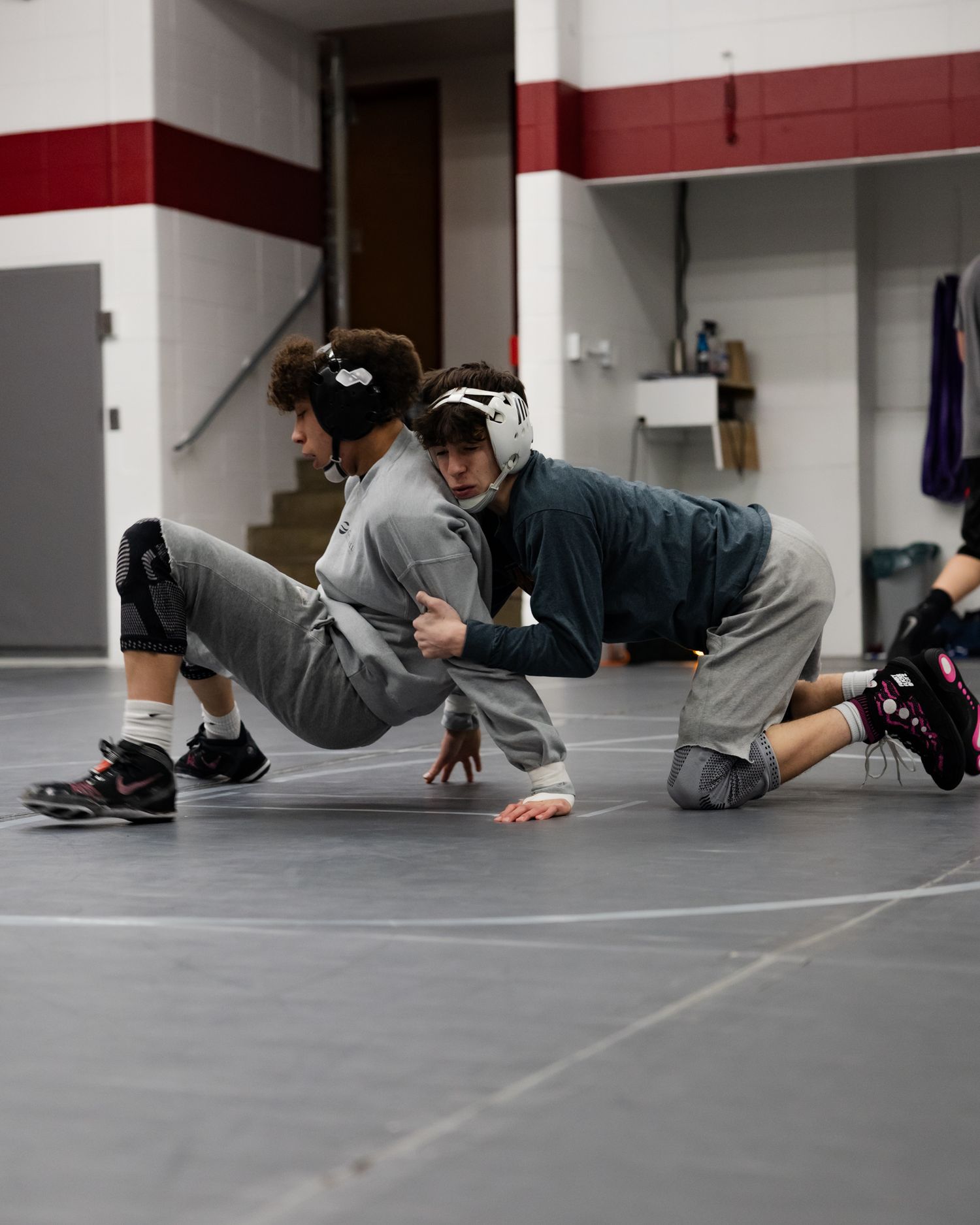 Two wrestlers in headgear practicing a takedown drill on a gray wrestling mat.