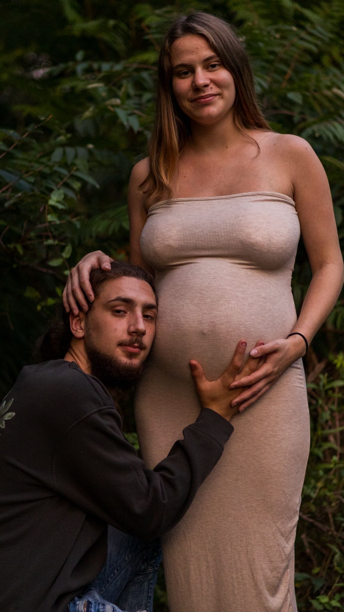 Couple poses outdoors for maternity photo, man kneeling beside pregnant woman in beige dress among green foliage.