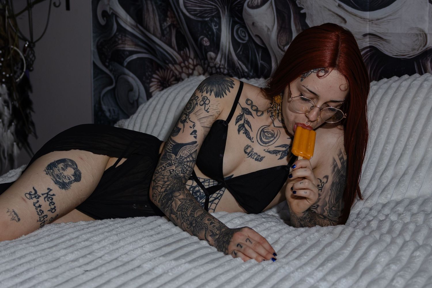 A heavily tattooed redhead woman in black outfit lies on a bed licking an orange popsicle.
