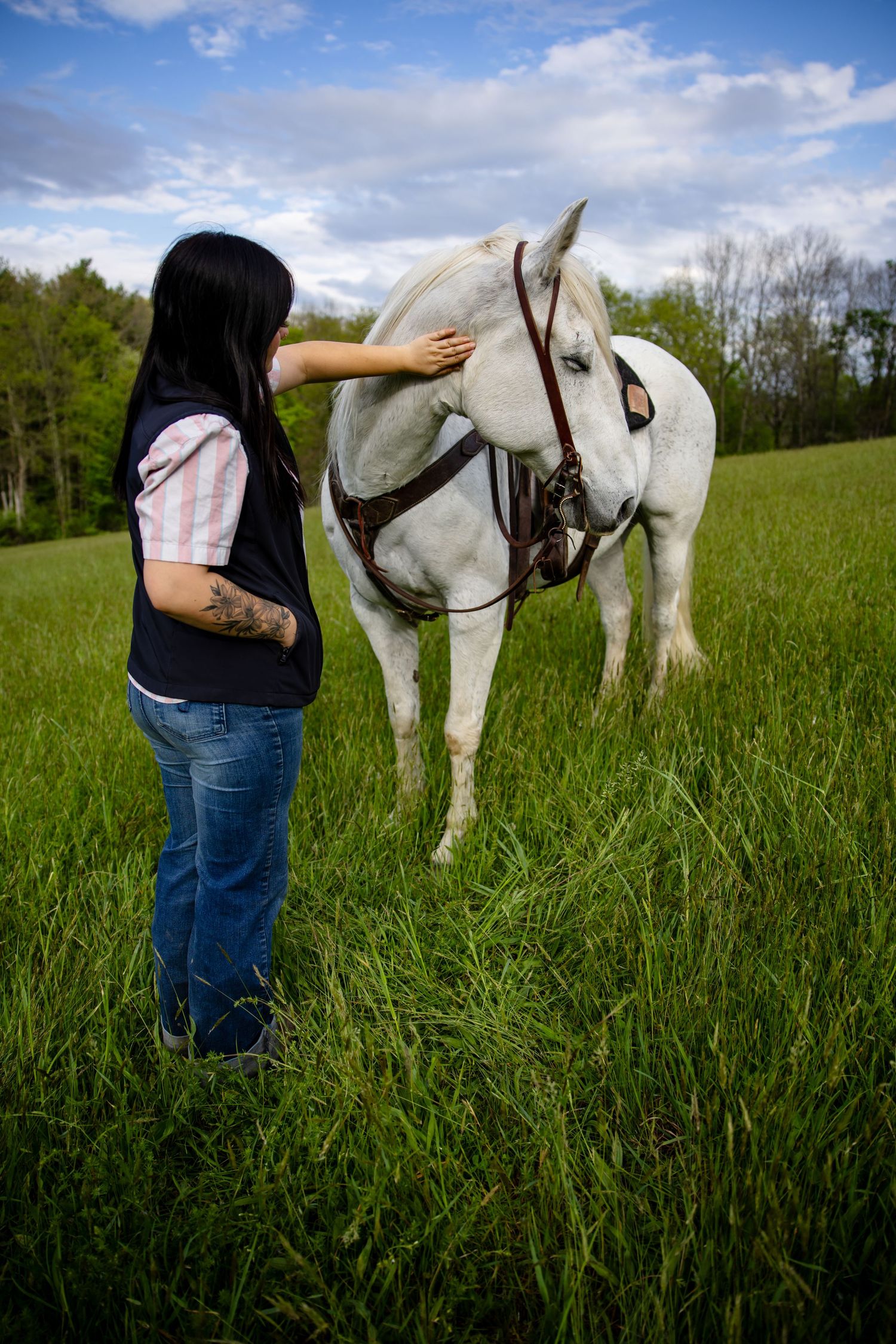Woman with tattoo petting a white horse wearing a bridle in a lush green field under a cloudy sky.