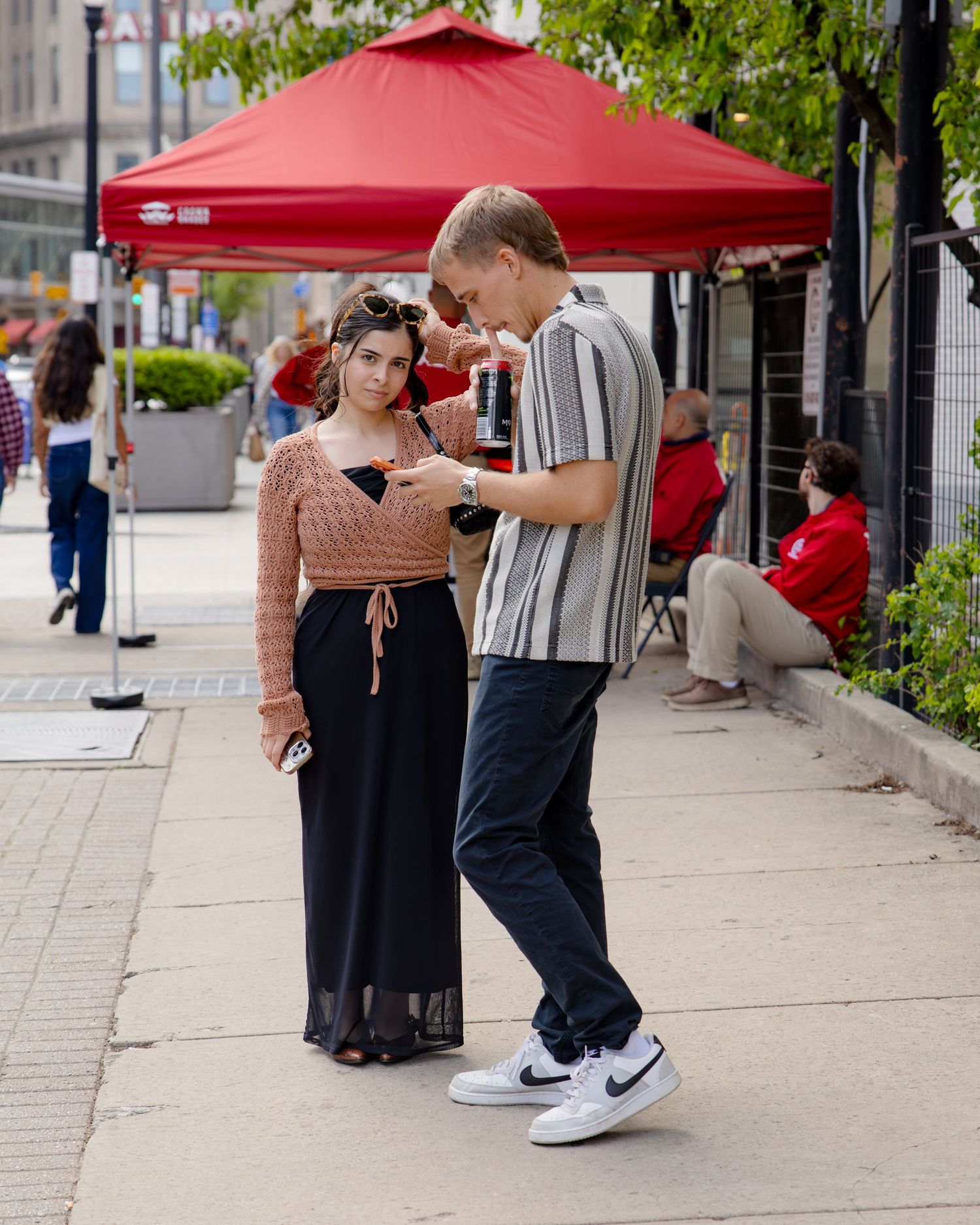 A young couple stands on a city sidewalk, the man showing something on his phone to the woman.