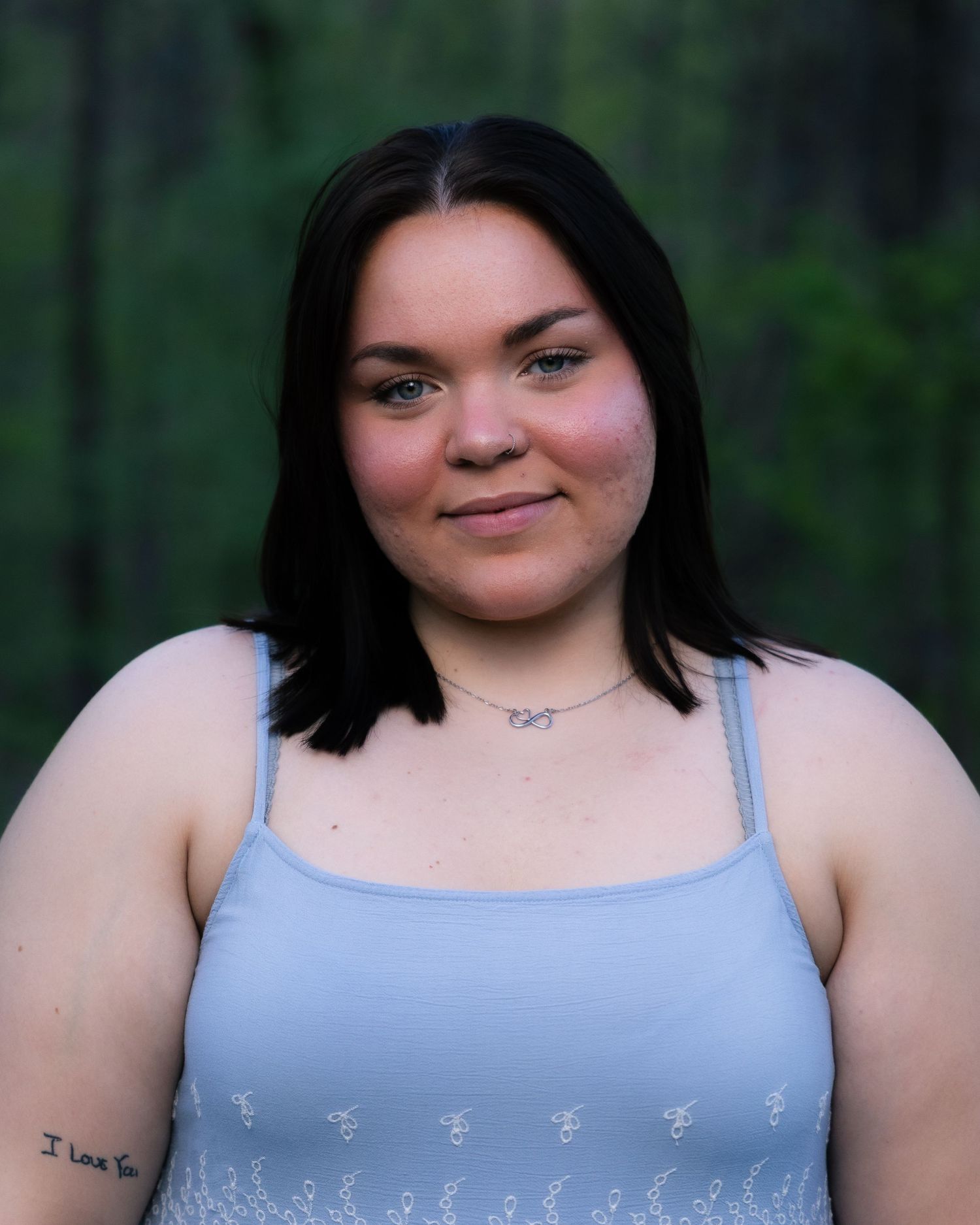 Young woman with dark hair wearing a light blue tank top, smiling outdoors with a forest background.