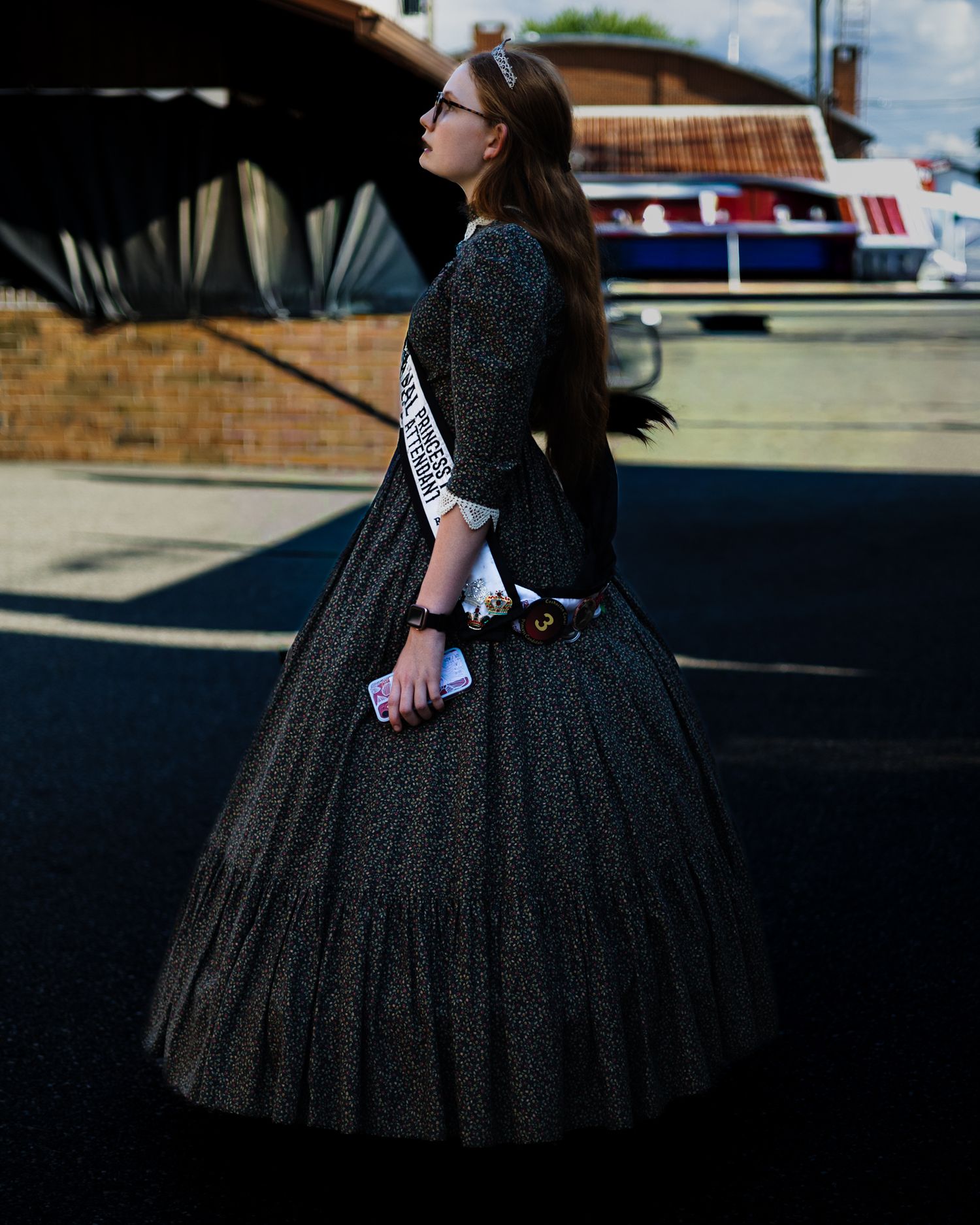 Young woman in a historic black ball gown with a pageant sash standing outdoors.