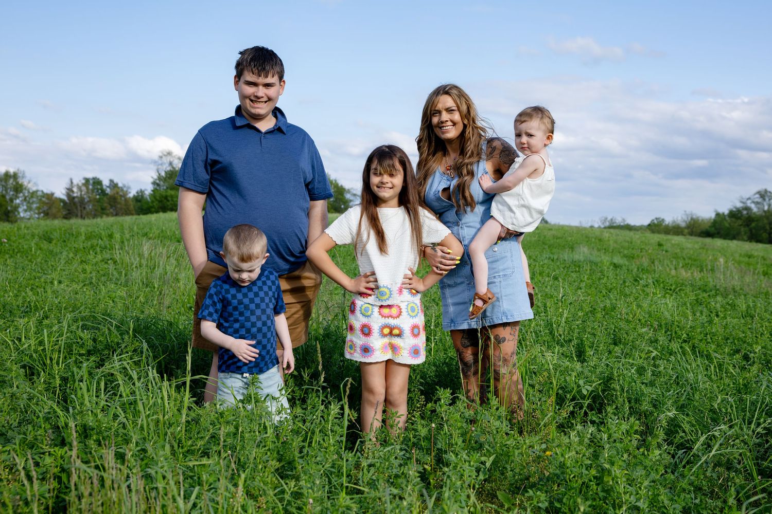 Family of five posing in a lush green field under a blue sky, with parents and three young children smiling.