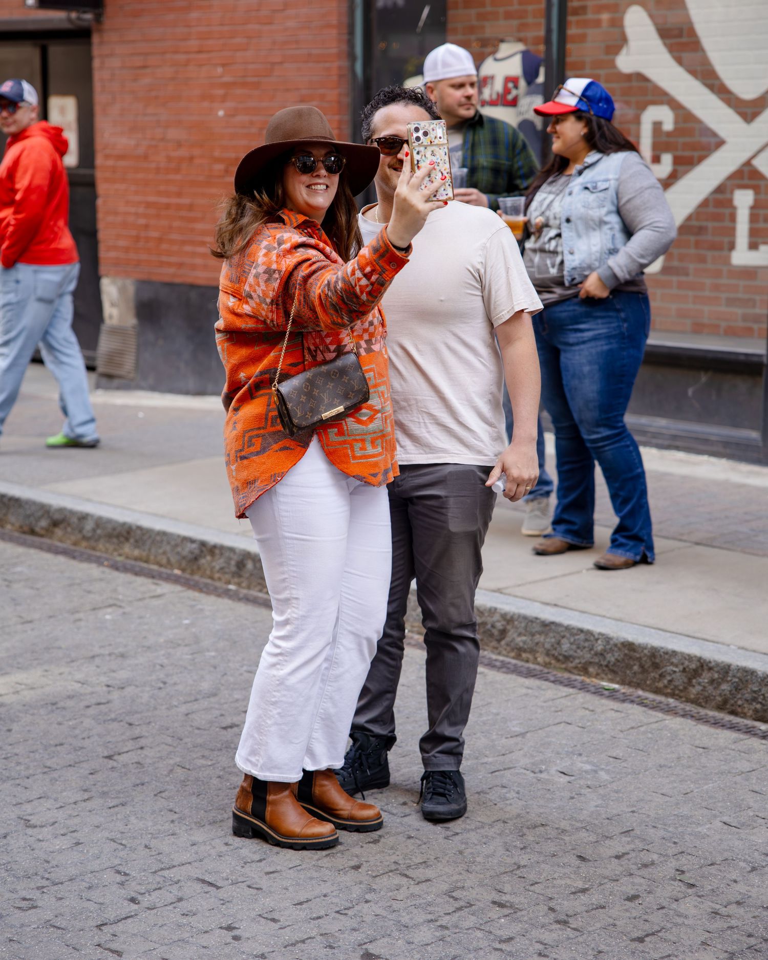 Couple taking a selfie on a brick street, woman wearing orange patterned jacket and brown hat.