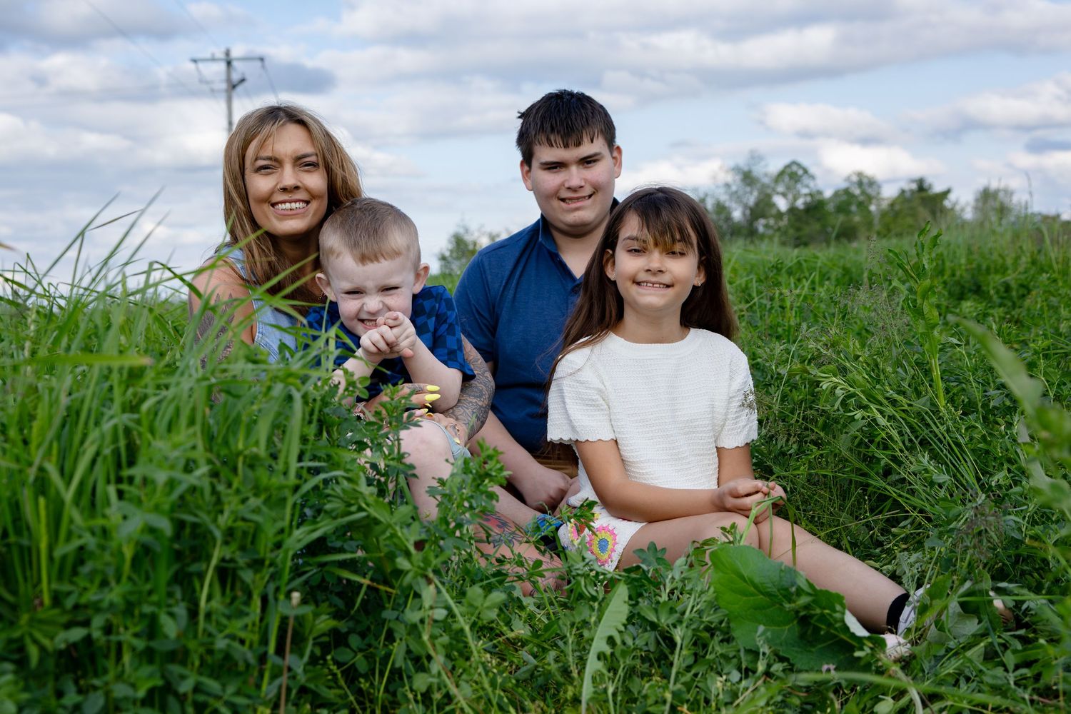 Family of four sitting together in lush green field, smiling outdoors on a cloudy day.