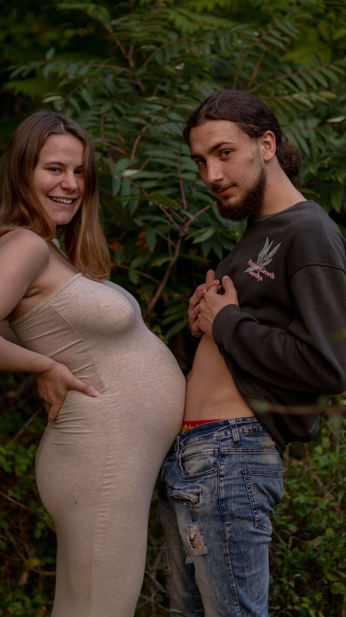 Smiling pregnant woman in beige dress poses with her partner outdoors in a green wooded setting.