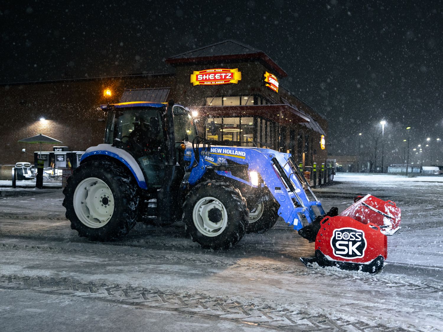 Blue tractor plowing snow in a Sheetz parking lot during a heavy nighttime snowstorm.