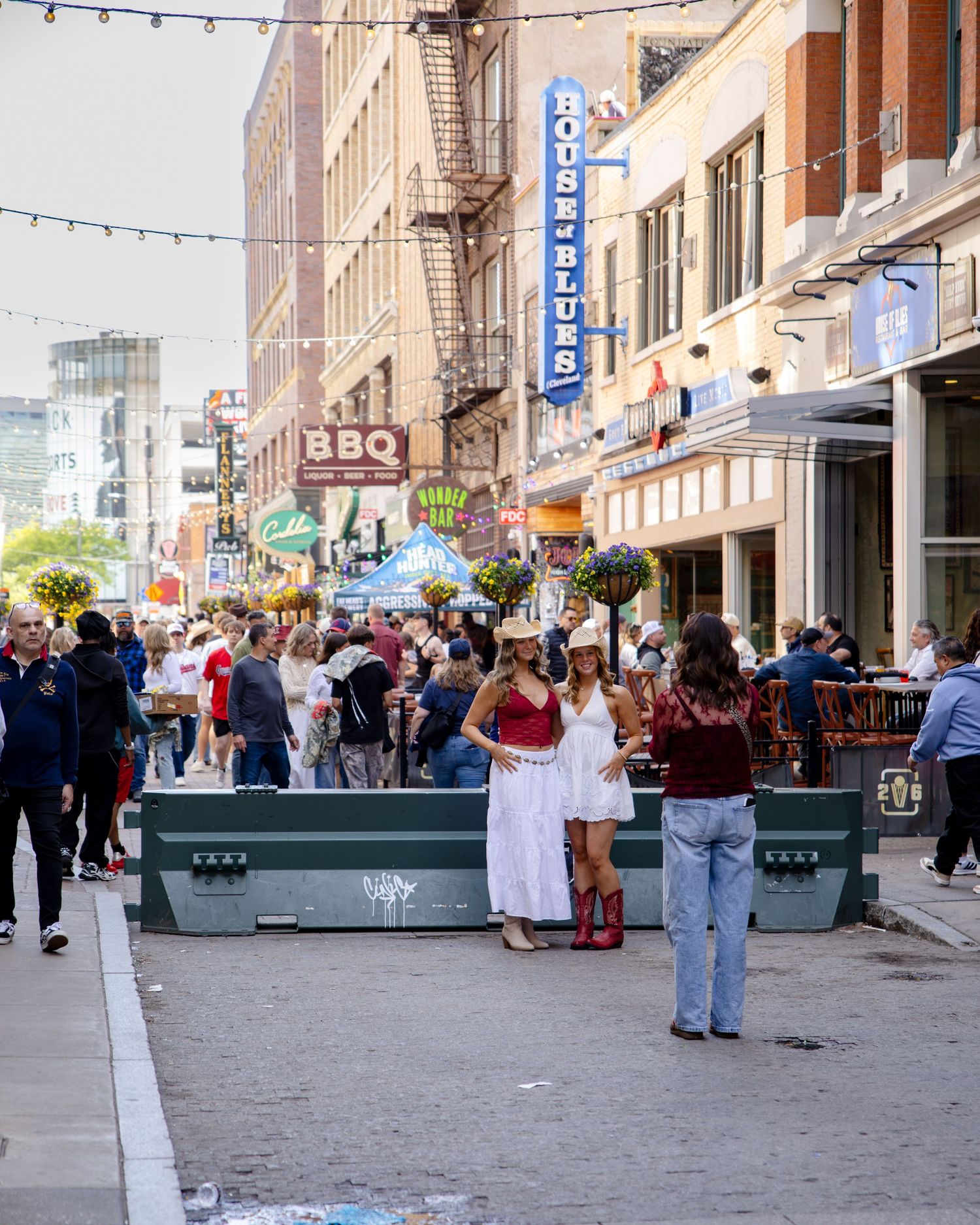 Busy street festival near House of Blues with crowds, outdoor dining, BBQ signs, and women in white dresses on a sunny day.