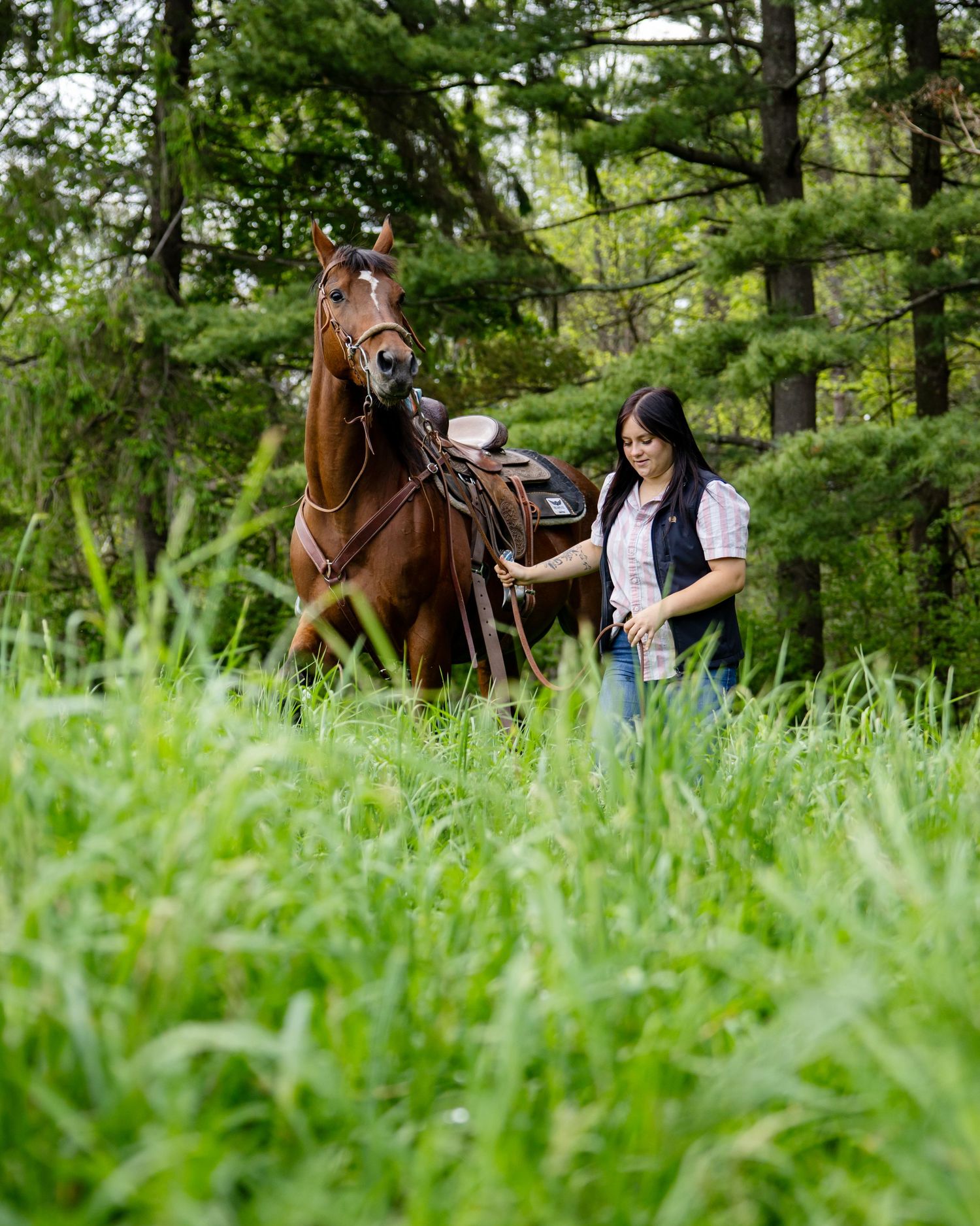 A woman leads a chestnut horse through lush green grass in a wooded setting.