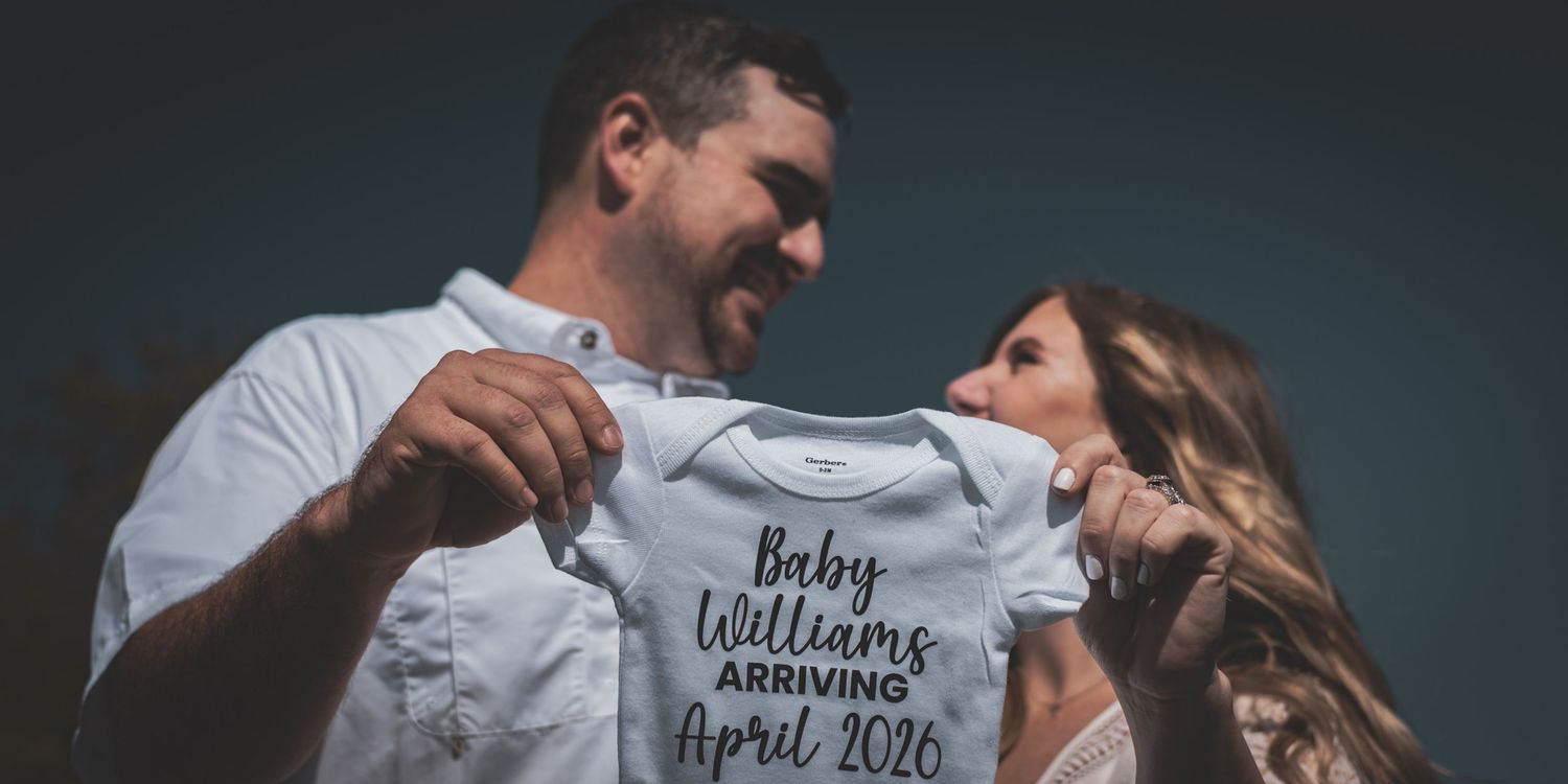 Couple holding a Baby Williams Arriving April 2026 onesie during maternity announcement.