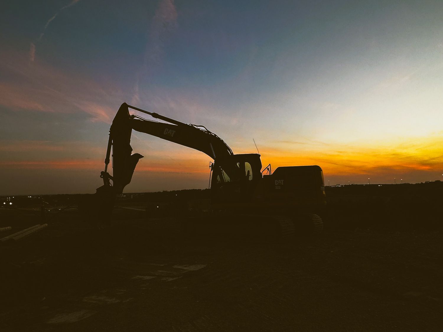 Excavator silhouette against a dramatic orange and blue sunset sky at a worksite.