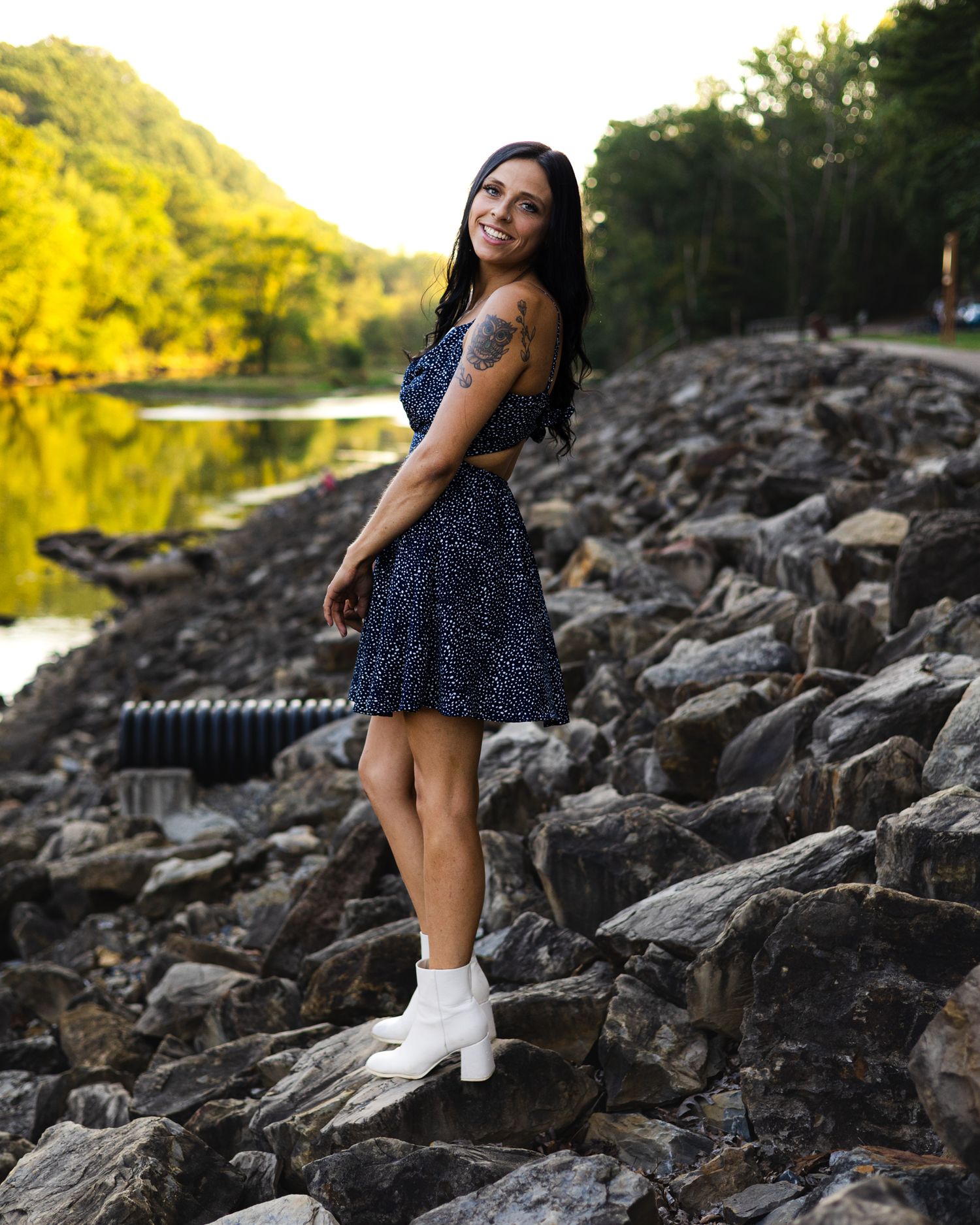 Brunette woman in blue floral dress and white boots posing on rocky riverbank.