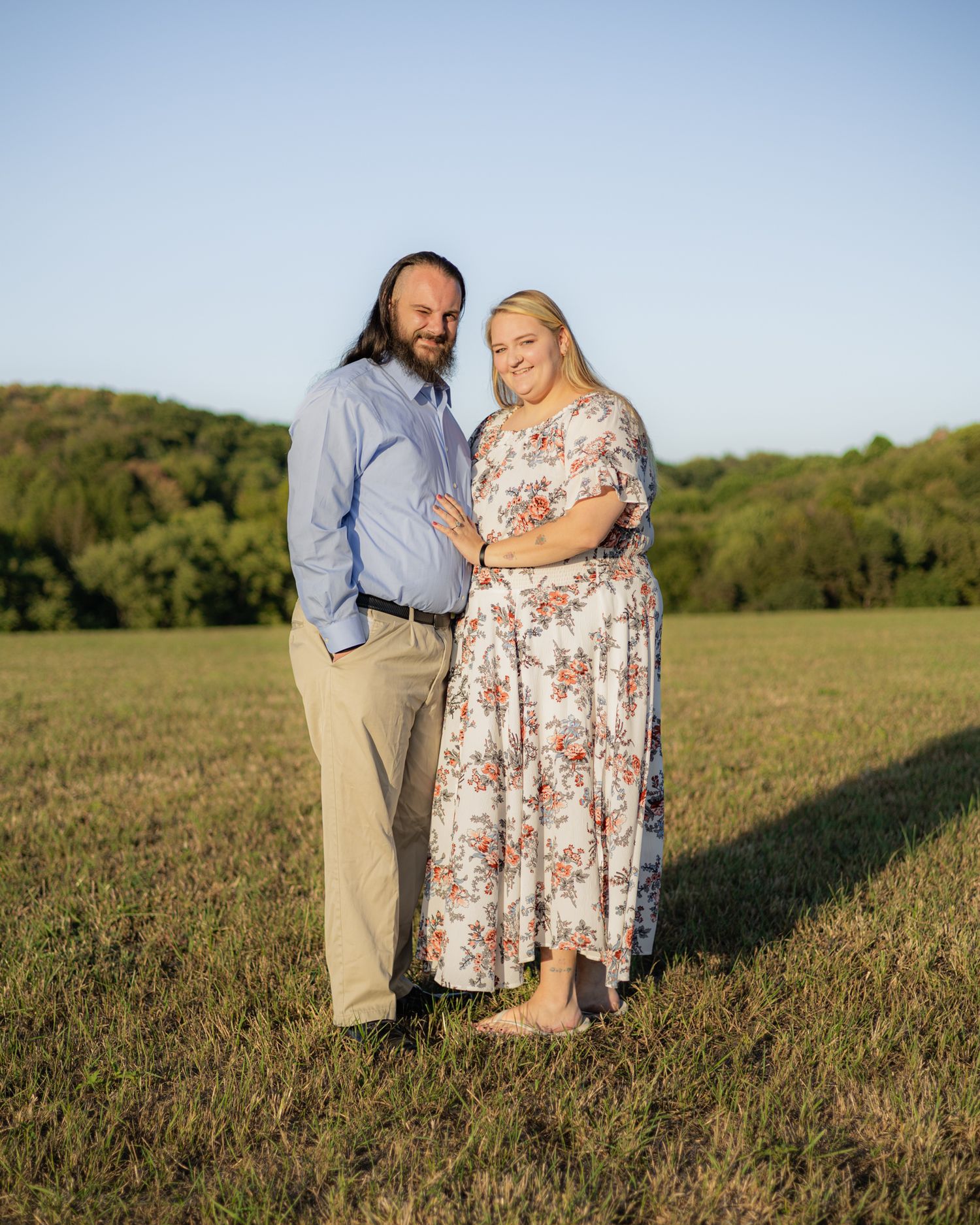 Couple posing together in a green field, man in blue shirt and woman in floral dress, with trees and blue sky behind.