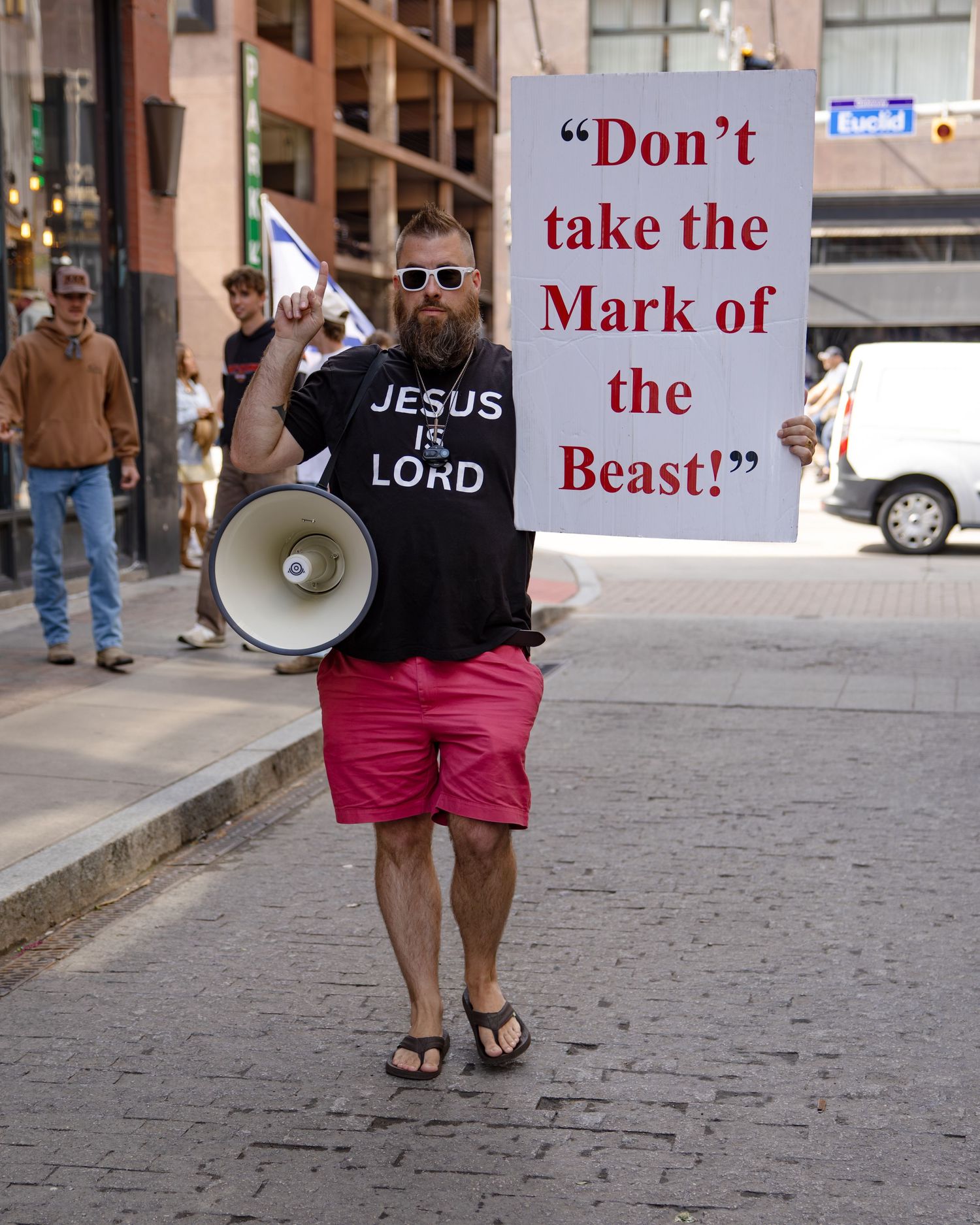Man with megaphone and Jesus is Lord shirt holds Don't Take the Mark of the Beast sign on city street.