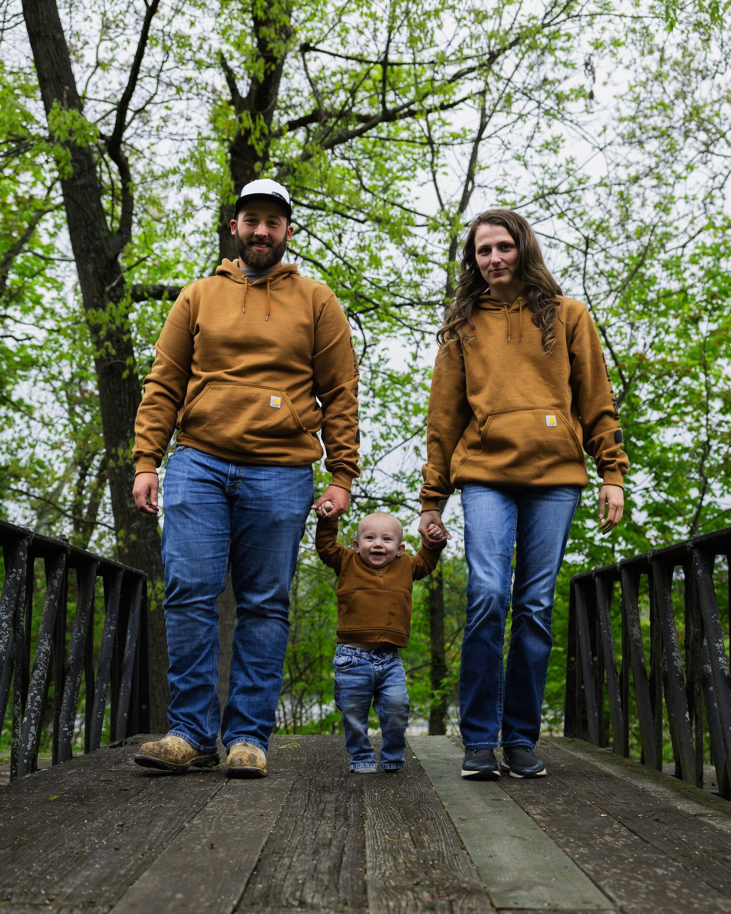 Family of three in matching brown Carhartt hoodies walks hand-in-hand on wooden bridge.