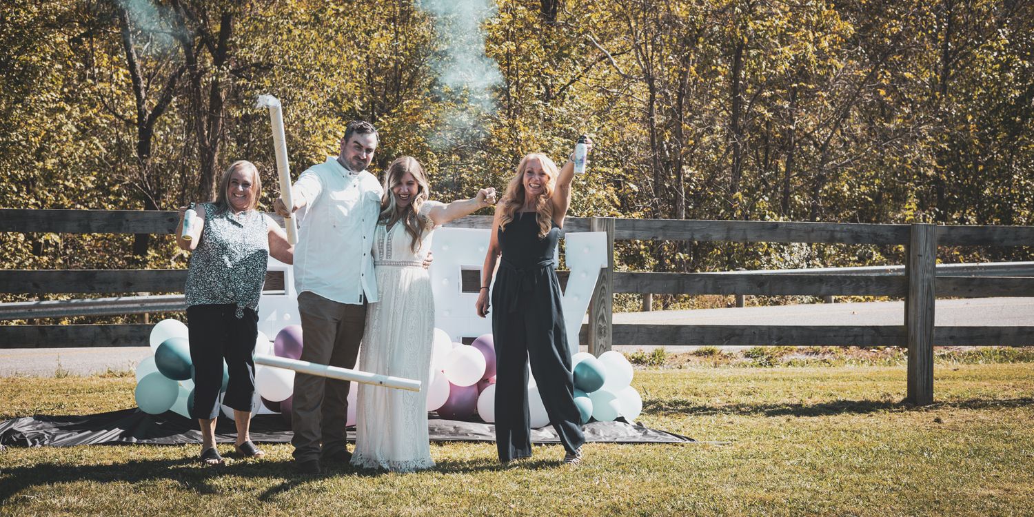 Family celebrating a gender reveal party outdoors with smoke cannon and colorful balloons.