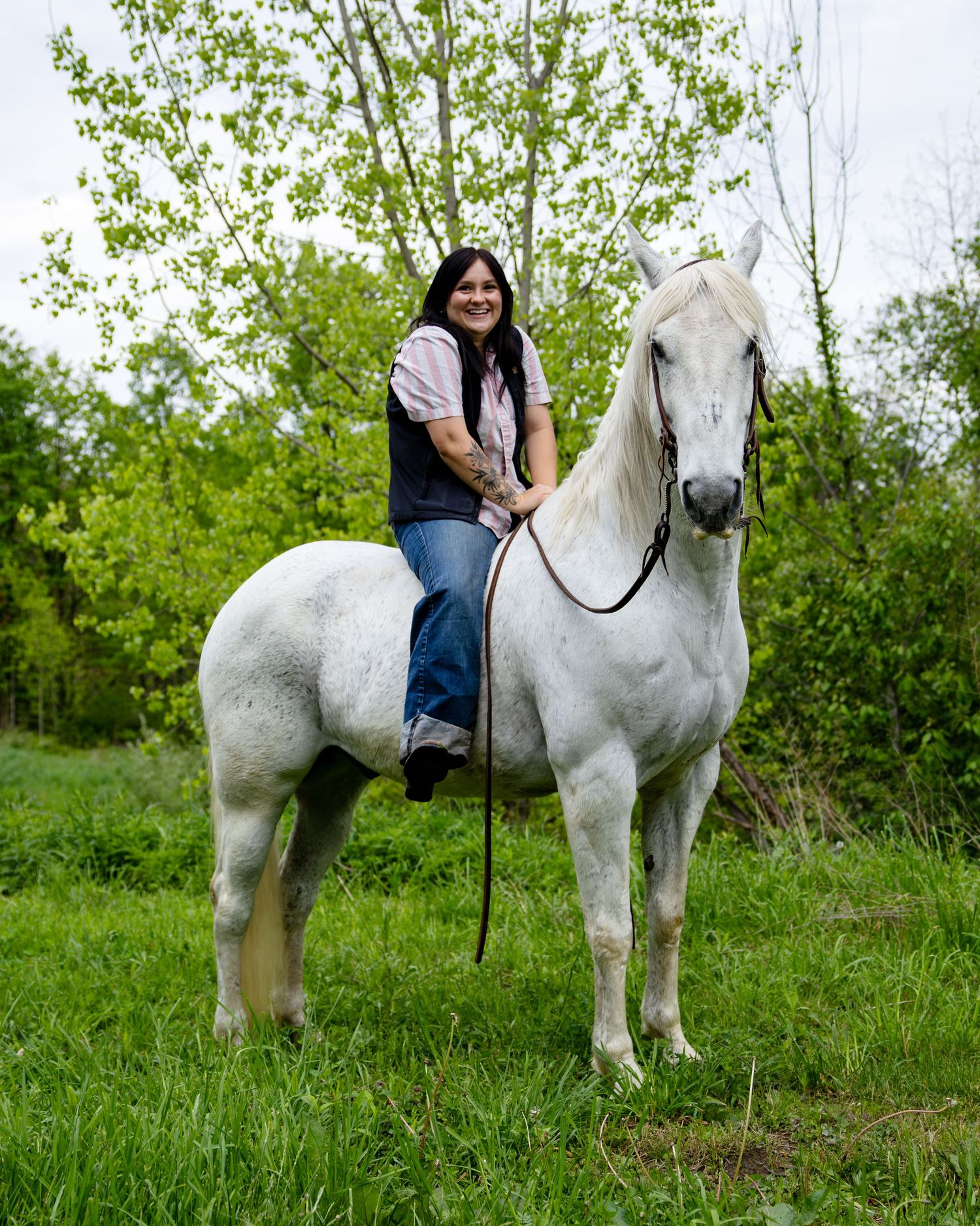 Woman smiling while riding a white horse in a lush green field surrounded by trees.