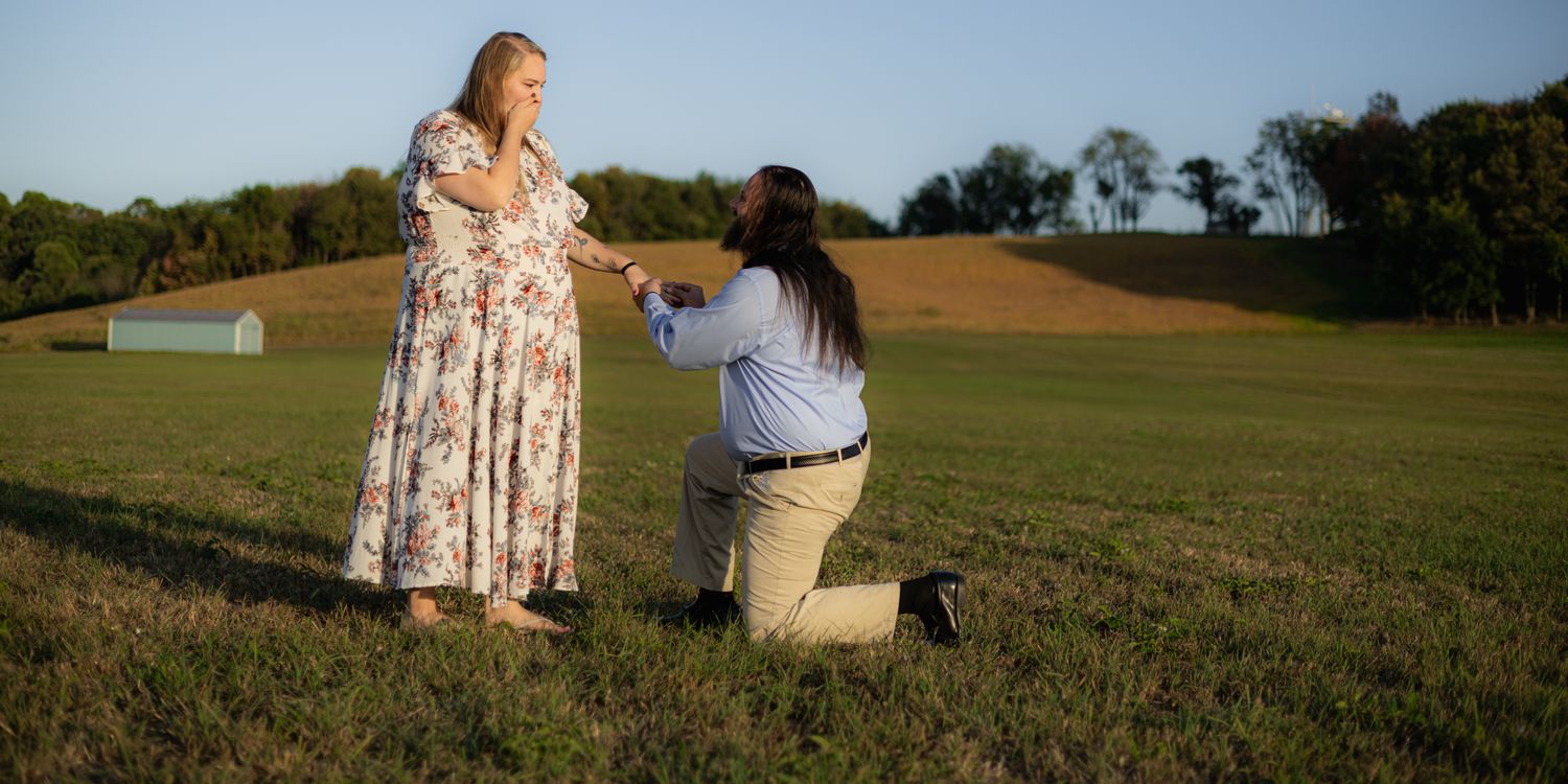 Person proposing on one knee in a green field to a woman in a floral dress at golden hour.
