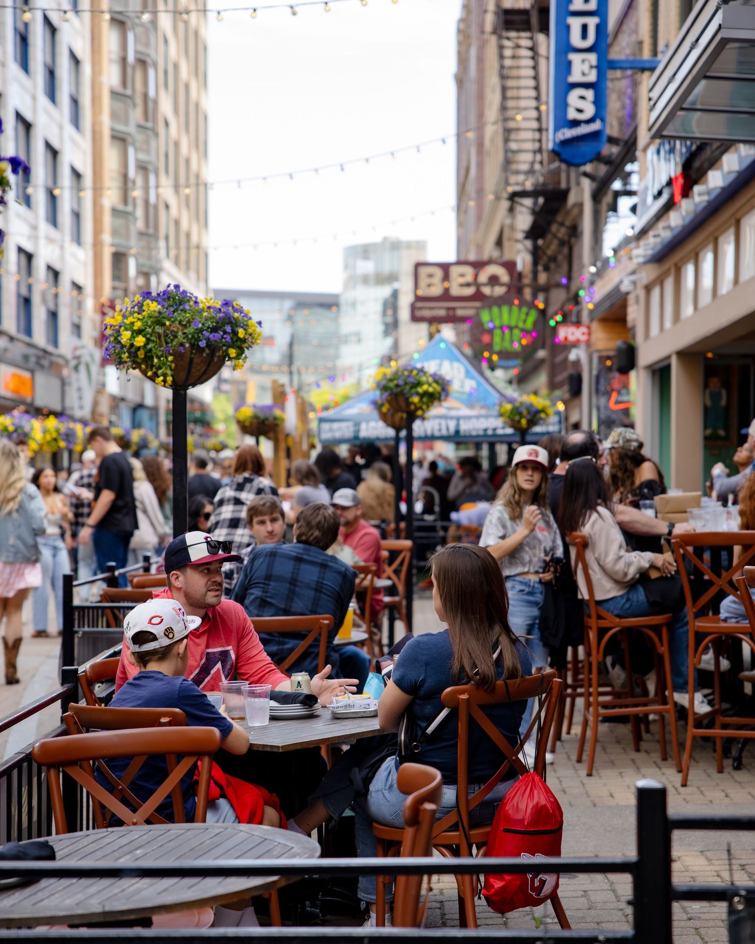 Busy outdoor patio dining on a vibrant city street lined with BBQ and blues restaurants, string lights, and flower baskets.