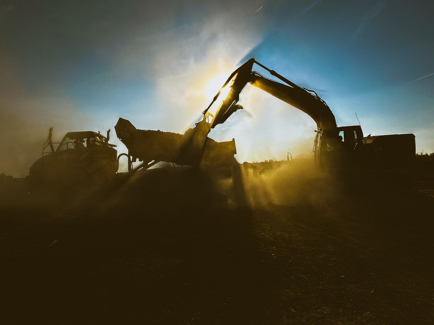 Excavator and bulldozer silhouetted against a dramatic golden sunset sky at a dusty construction site.