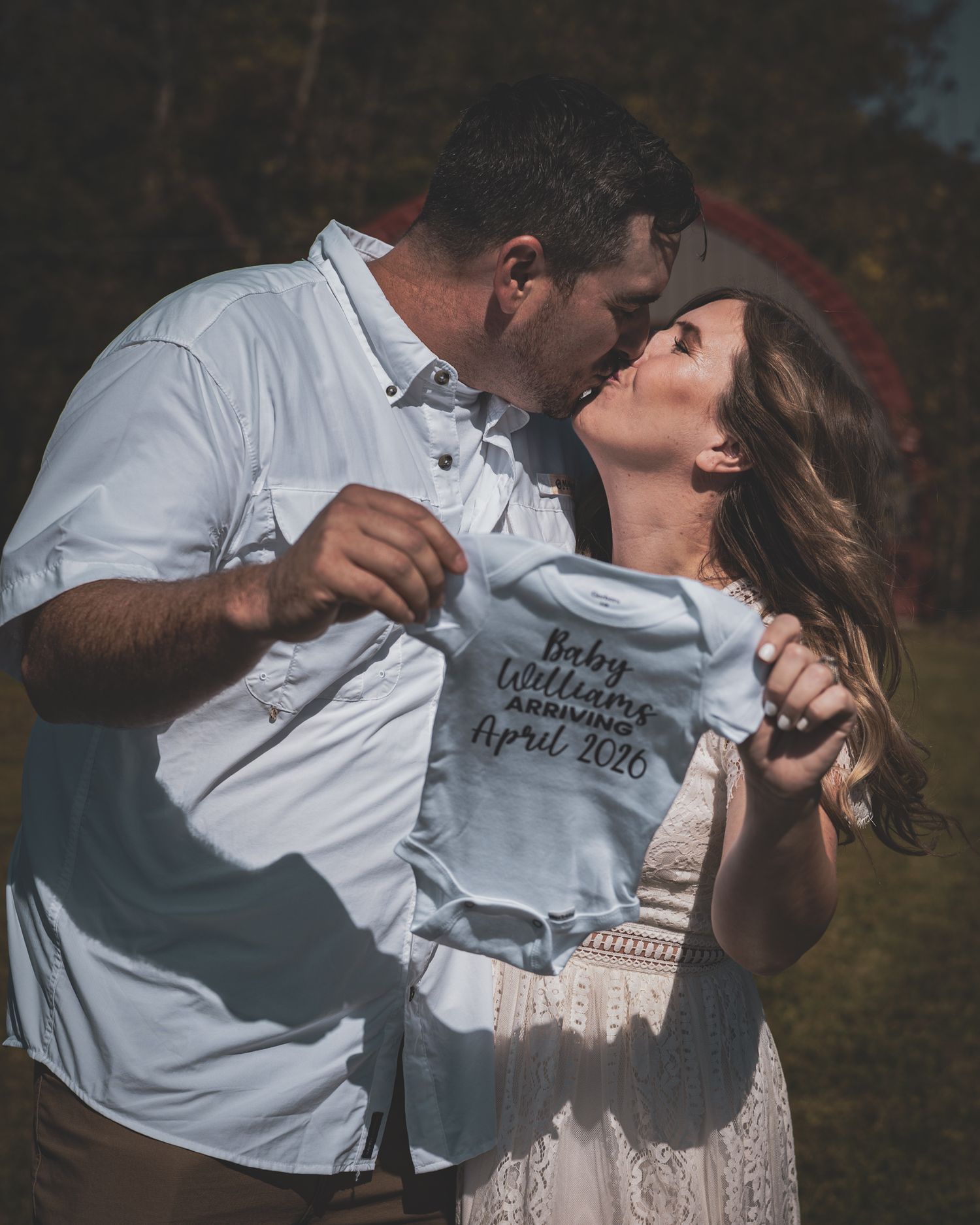 Couple kissing while holding Baby Williams Arriving April 2026 onesie during maternity shoot.