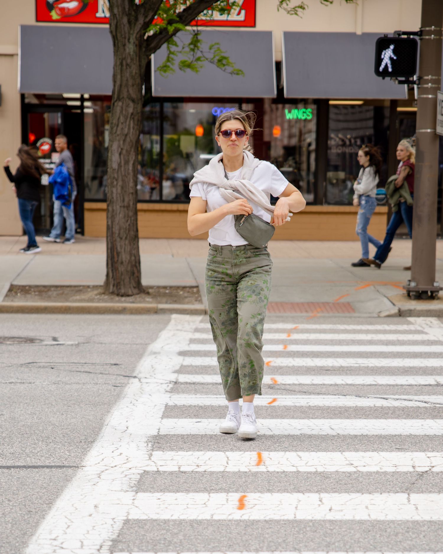 Woman in camo pants and white tee crosses a city crosswalk carrying a gray bag, wearing white sneakers.