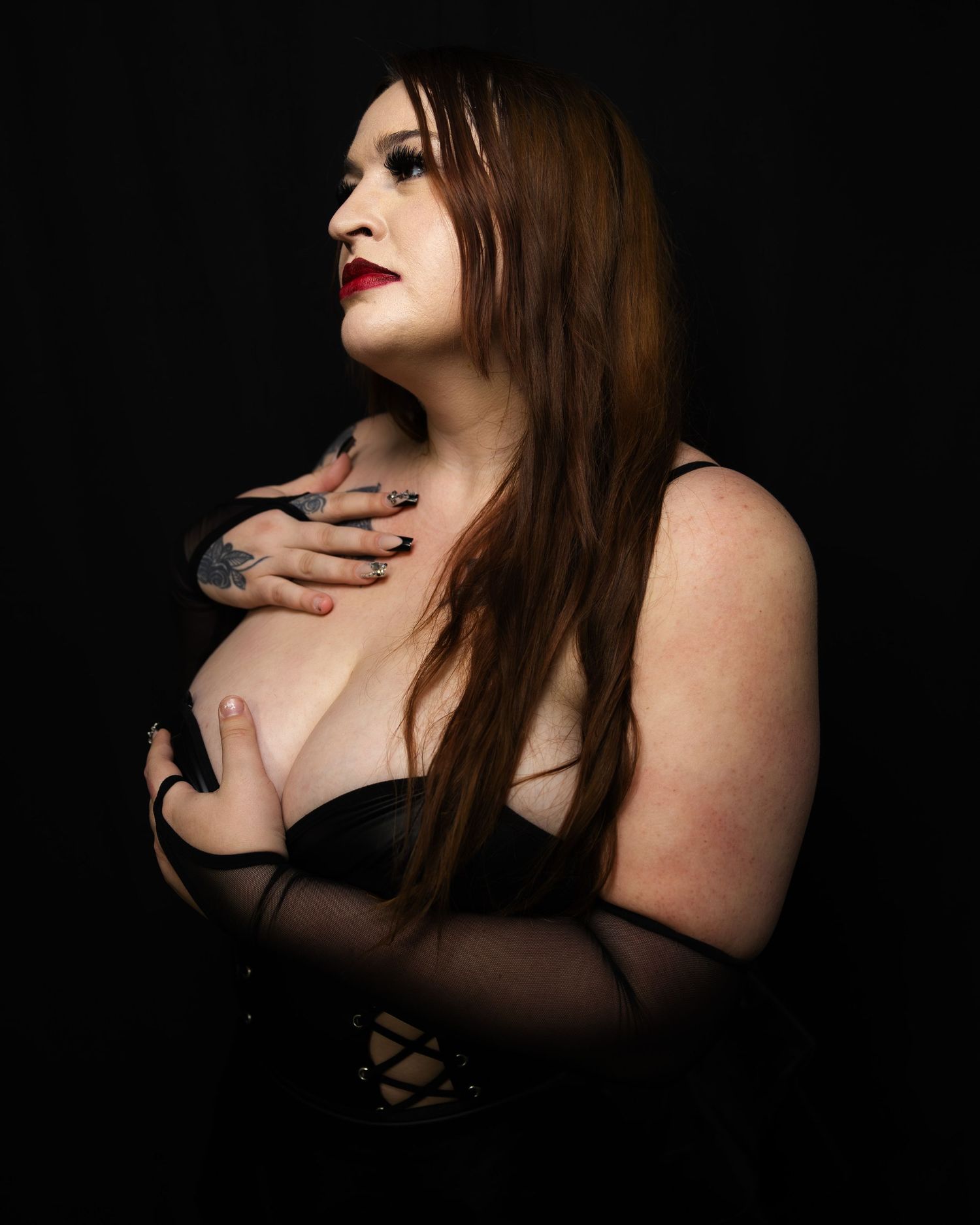 A tattooed redhead woman in black lingerie poses against a dark background with bold red lips.
