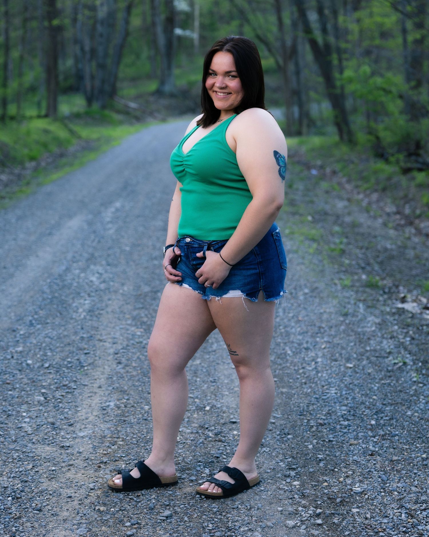 Smiling woman in green tank top and denim shorts standing on forest gravel road.