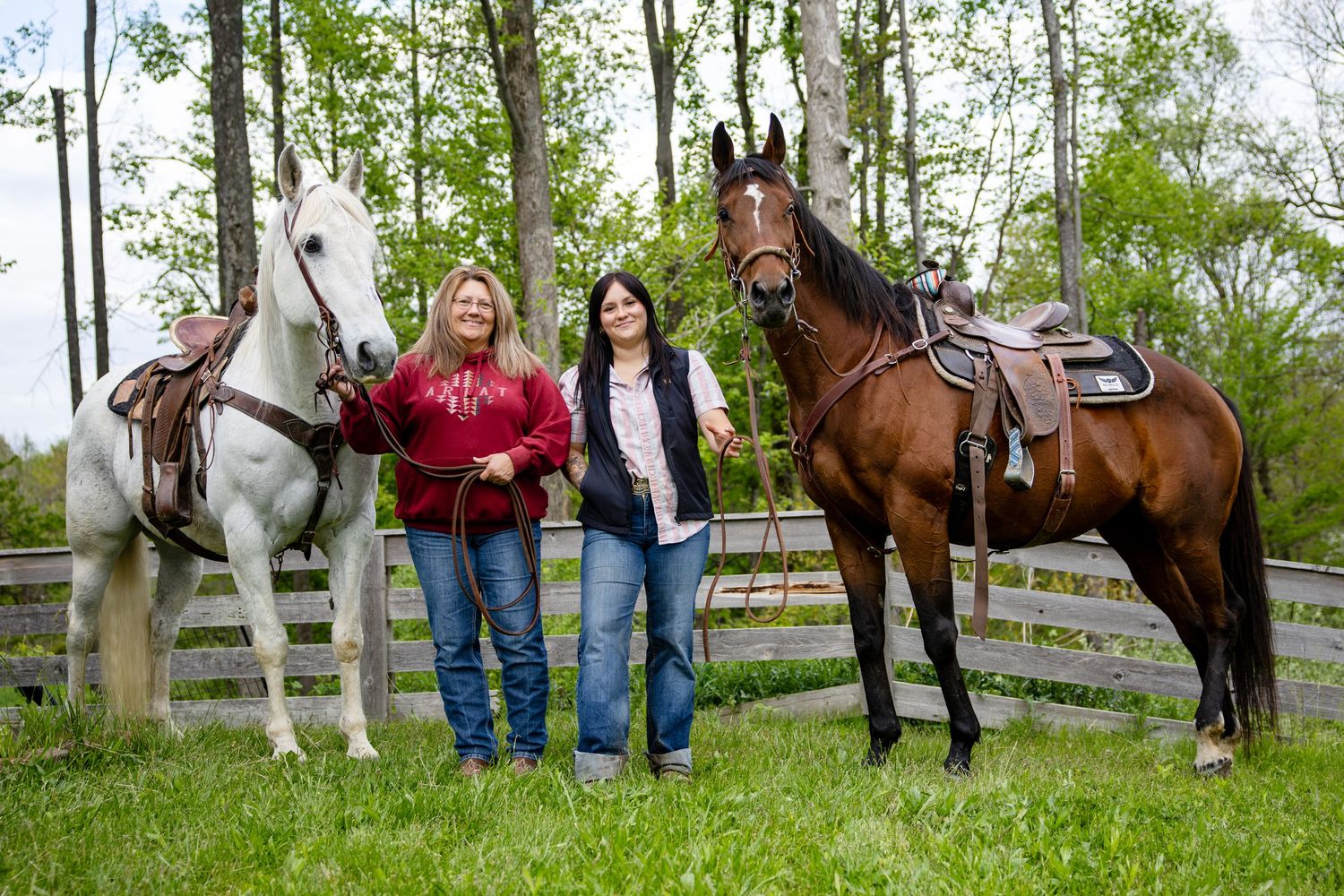 Two women standing outdoors with a white and a brown saddled horse on green grass near a wooden fence.
