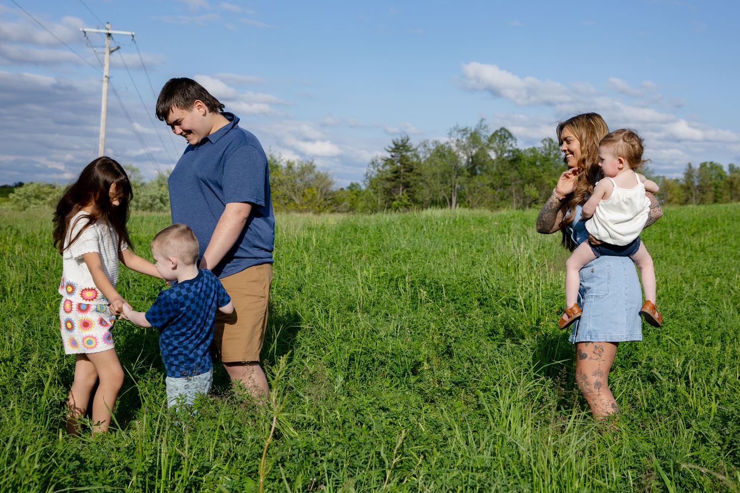 Family of four with young children enjoying a sunny day in a lush green field under a blue sky.