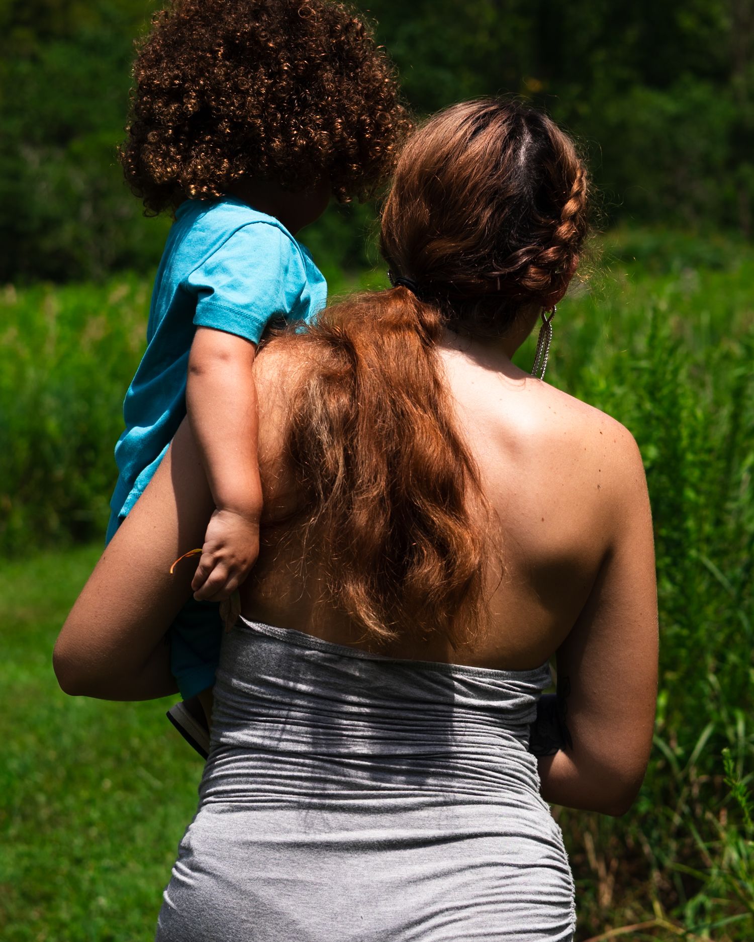 Mother in gray strapless dress carrying young child outdoors in green setting.