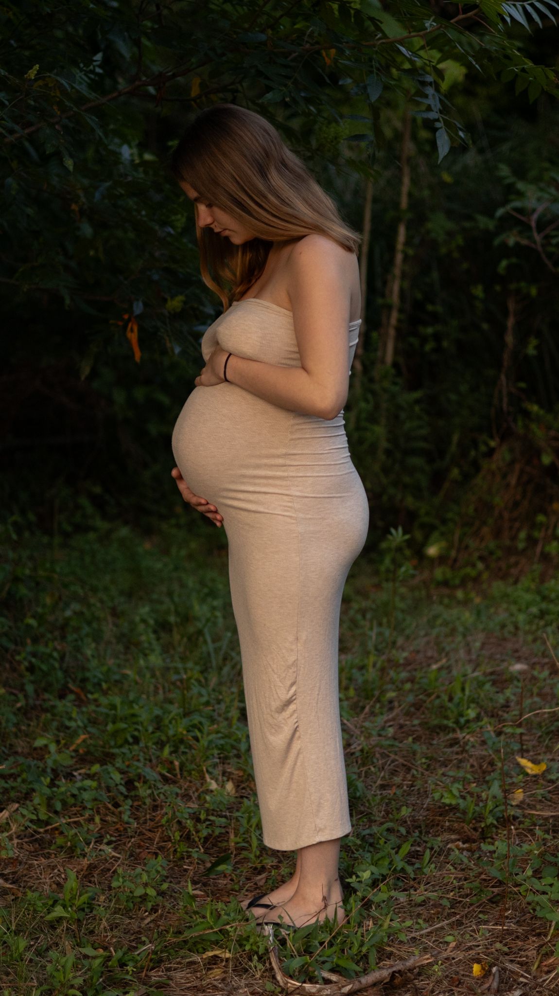 Pregnant woman in beige tube dress cradles her baby bump while standing in a natural outdoor setting.