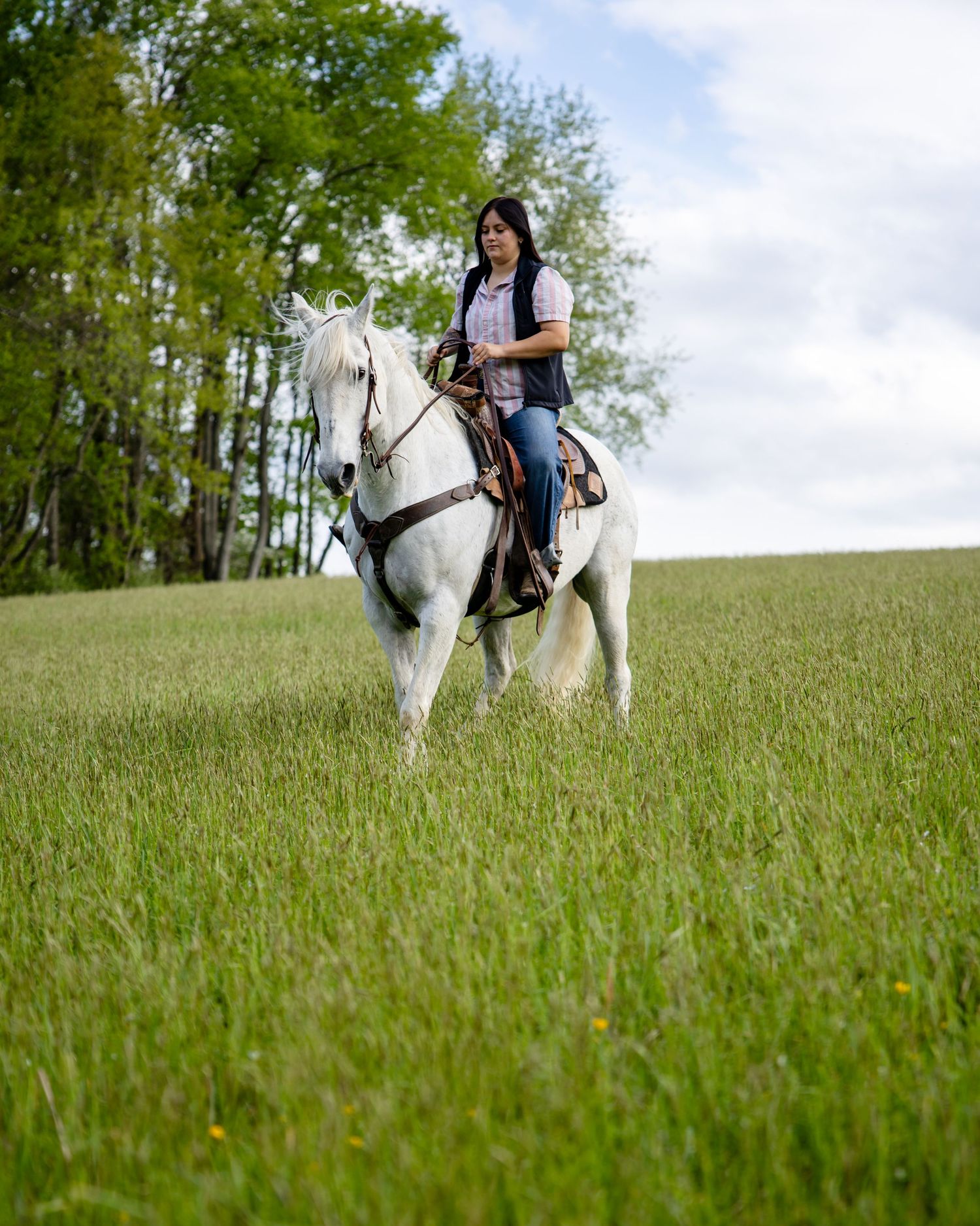 Woman riding a white horse through a lush green field with trees in the background on a cloudy day.
