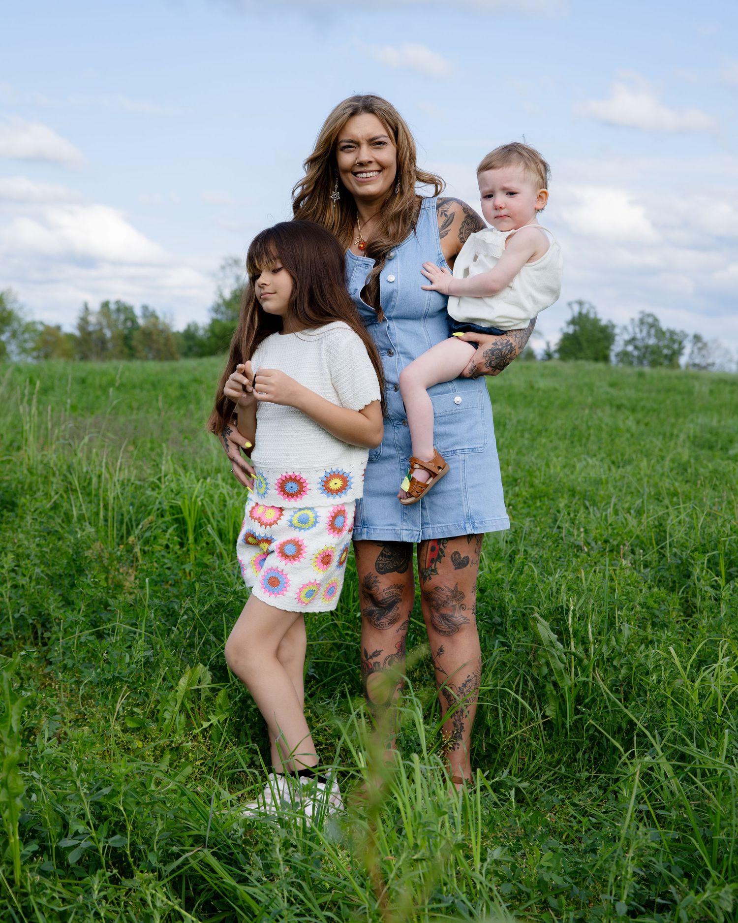 Tattooed mother holding baby stands with young daughter in grassy field, wearing denim dress and crochet skirt.