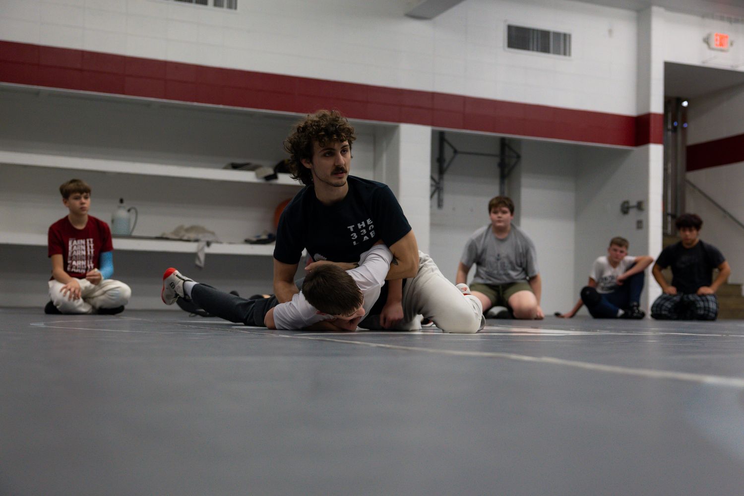 Wrestling coach demonstrating a ground hold technique to students on a gym mat.