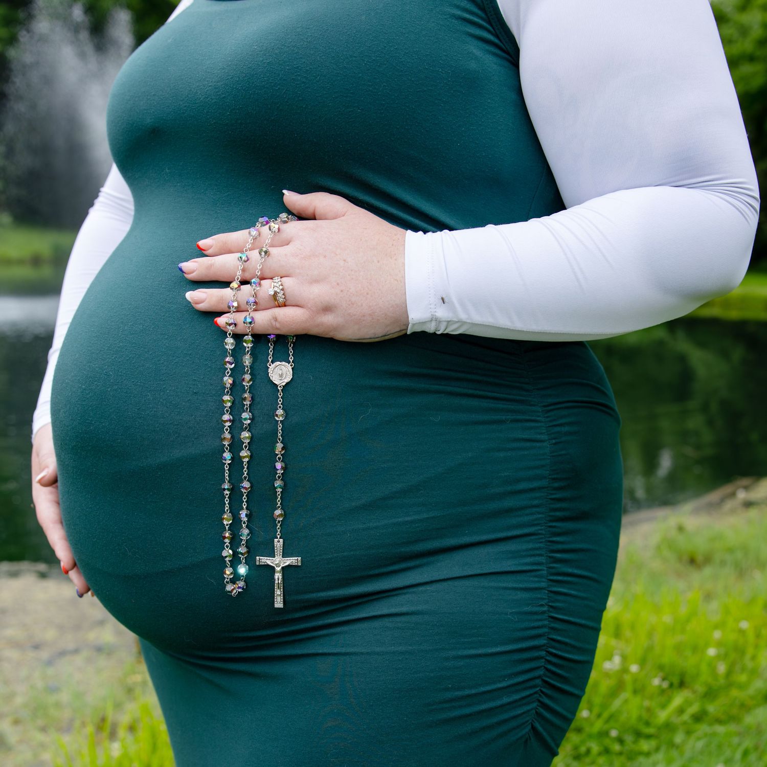 Pregnant woman in green dress holding rosary beads, showcasing maternity fashion outdoors.