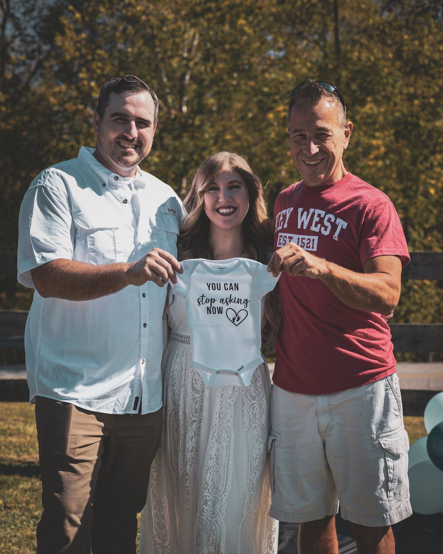 Two men and a pregnant woman smile holding a You Can Stop Asking Now baby onesie at a gender reveal.