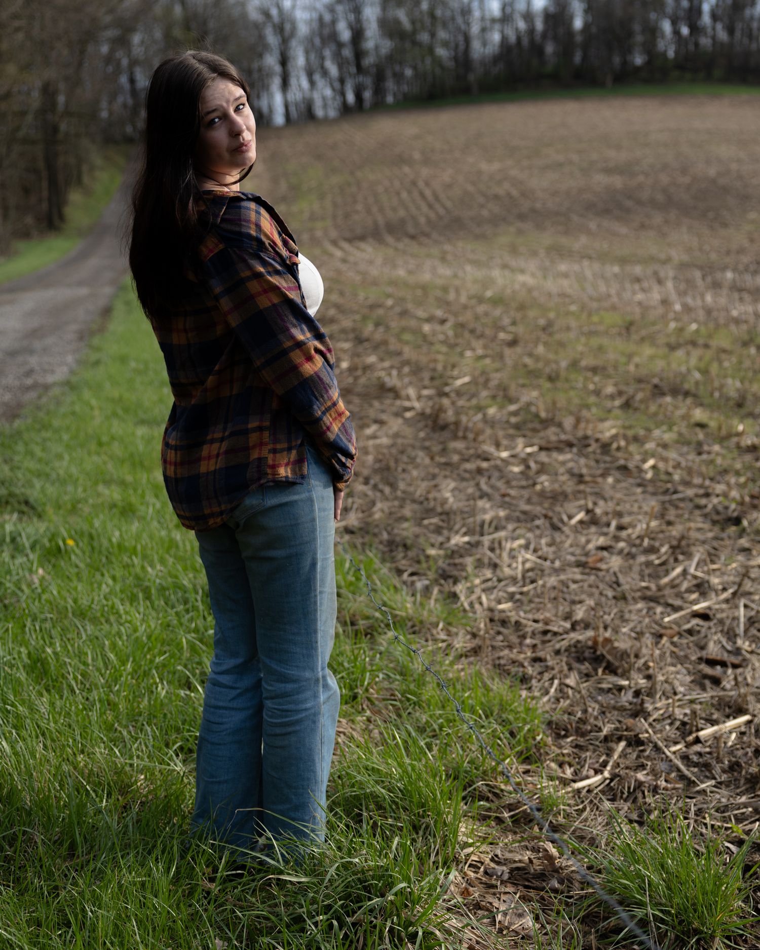 Young woman in plaid shirt and jeans standing beside rural countryside field.