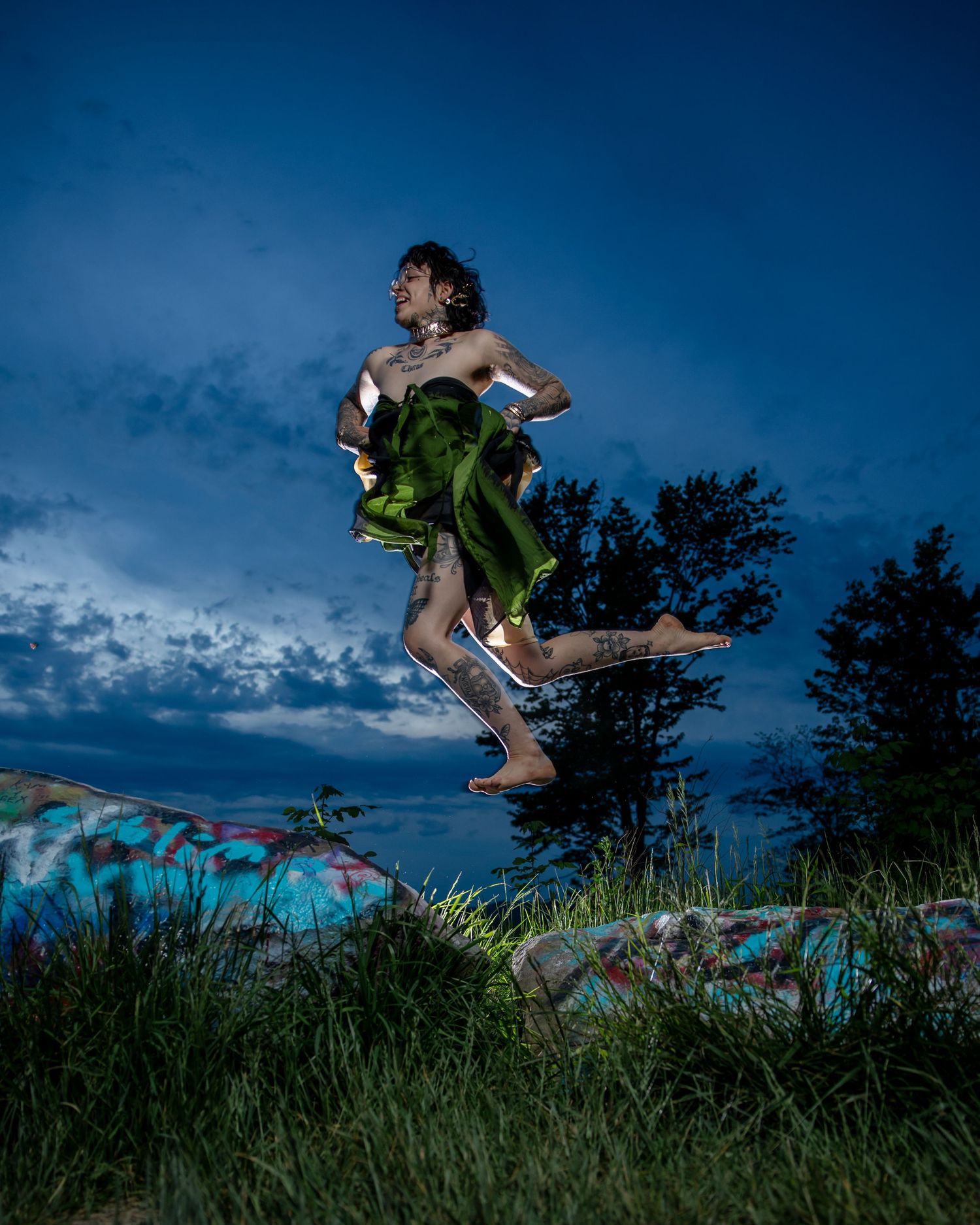 A tattooed person leaps mid-air outdoors at dusk, draped in green fabric, above graffiti-covered surfaces with trees and blue sky.