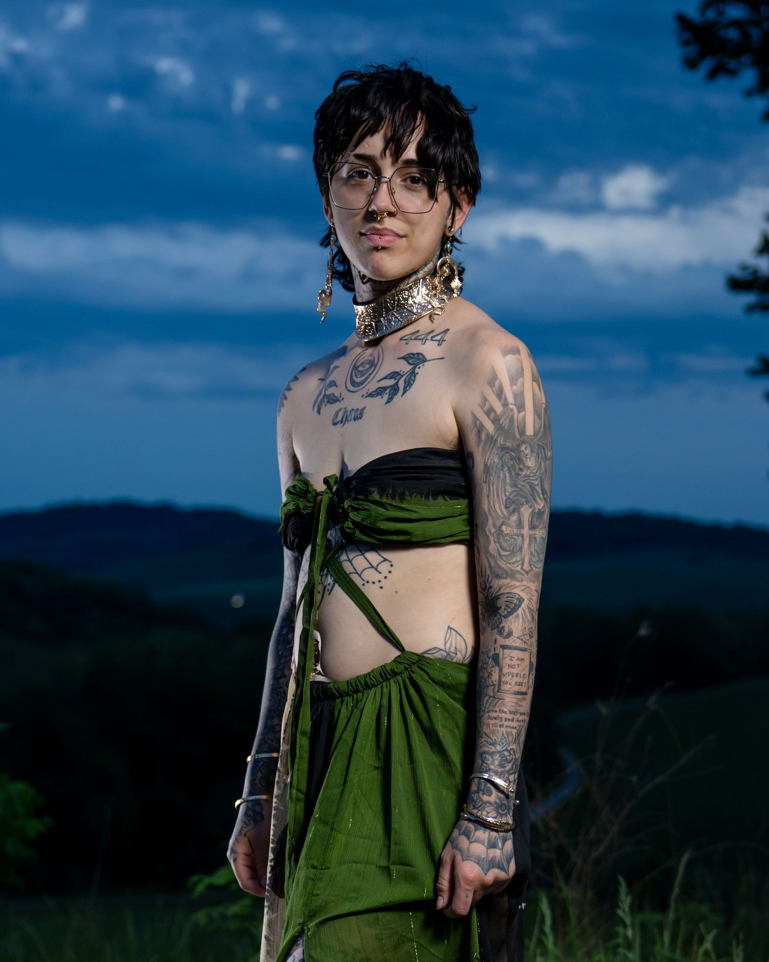 Tattooed person wearing green wrap skirt and crop top poses outdoors at dusk with hills in background.