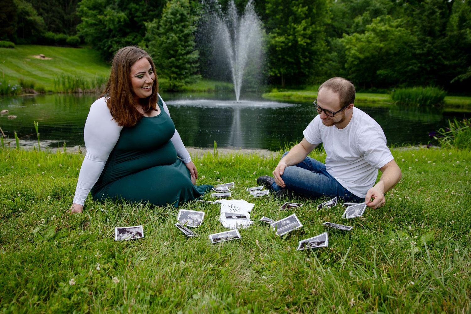 Pregnant couple sitting on grass by a pond with fountain, looking at ultrasound photos spread out between them.