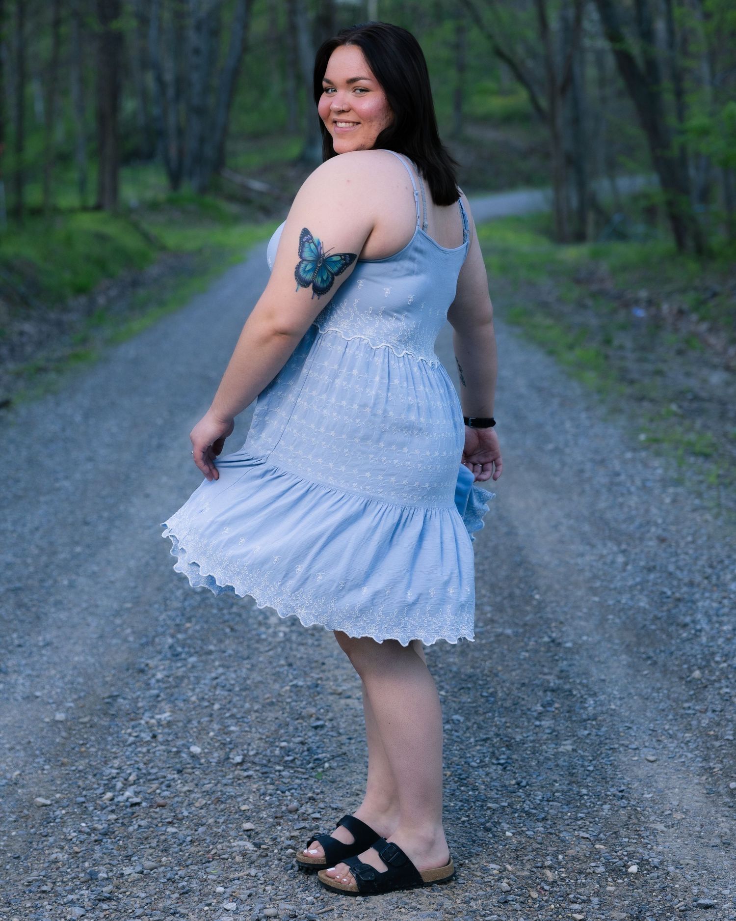 Woman in light blue sundress with butterfly tattoo posing on wooded gravel path.
